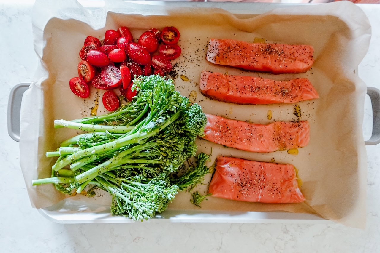 A baking tray lined with parchment paper holds four raw salmon fillets, a bunch of broccolini, and sliced cherry tomatoes, all seasoned with pepper and ready to be cooked.