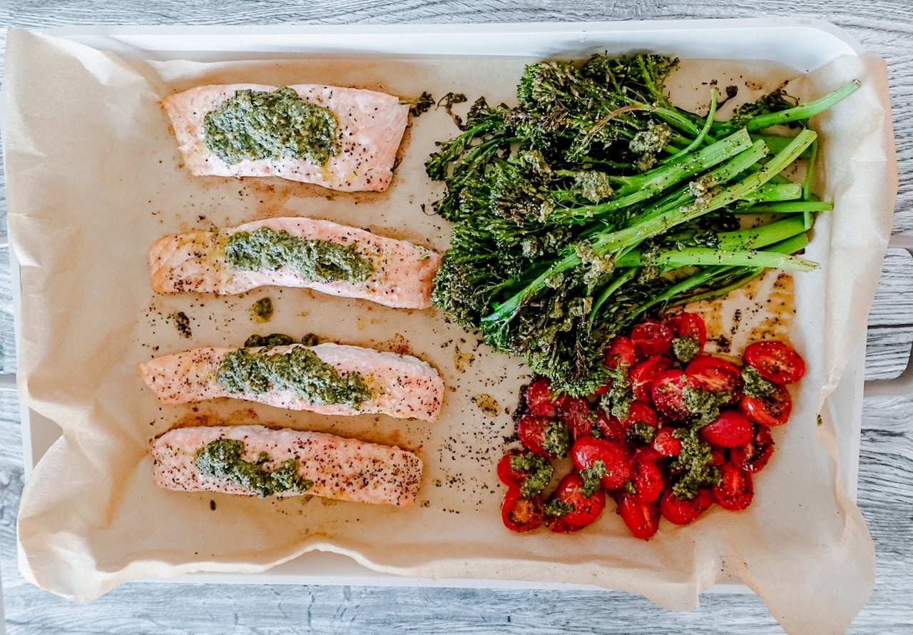 Four salmon fillets topped with green pesto, roasted broccolini, and halved cherry tomatoes arranged on a parchment-lined baking tray. The food is seasoned with herbs and spices.