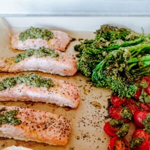 Baked salmon fillets topped with green pesto sit on a parchment-lined tray alongside roasted broccolini and cherry tomatoes, all seasoned with pepper and herbs.