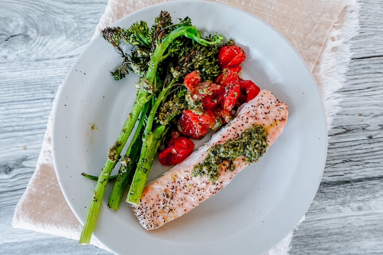 A plate with a baked salmon fillet topped with green pesto, served alongside roasted broccolini and cherry tomatoes on a light-colored napkin and wooden background.