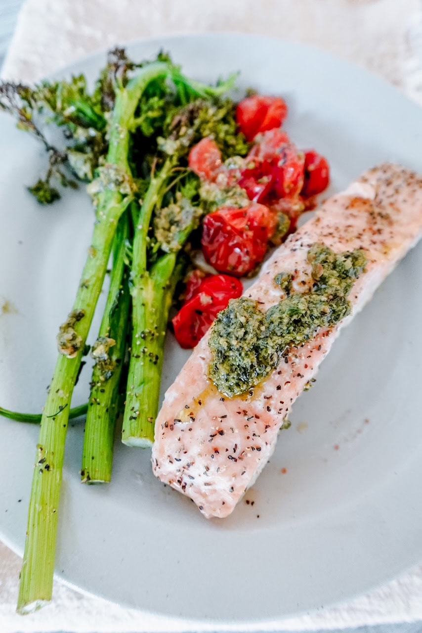 A plate with baked salmon topped with green pesto, roasted cherry tomatoes, and broccolini, served on a light-colored dish.
