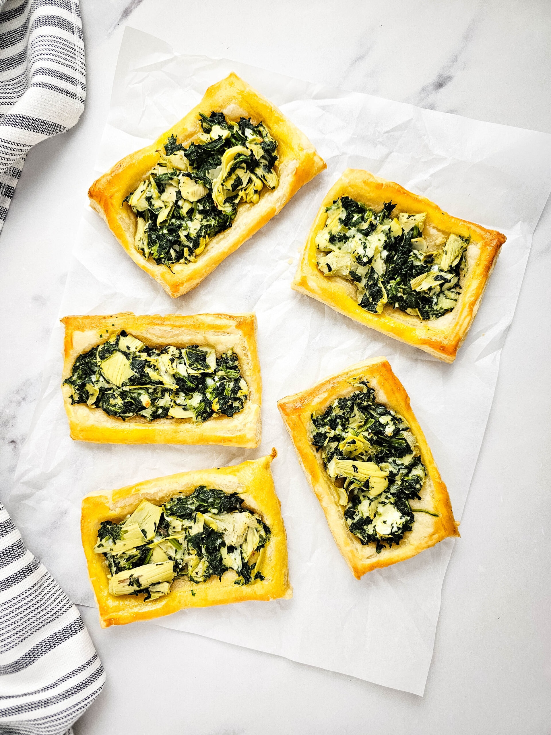 An overhead view of five rectangular puff pastry tarts topped with spinach, artichokes, and cheese are arranged on a sheet of parchment paper on a marble surface. A striped cloth napkin is at the left side of the shot.