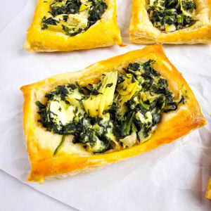 Three rectangular puff pastry tarts topped with a mixture of spinach, artichokes, and cheese, arranged on white parchment paper.