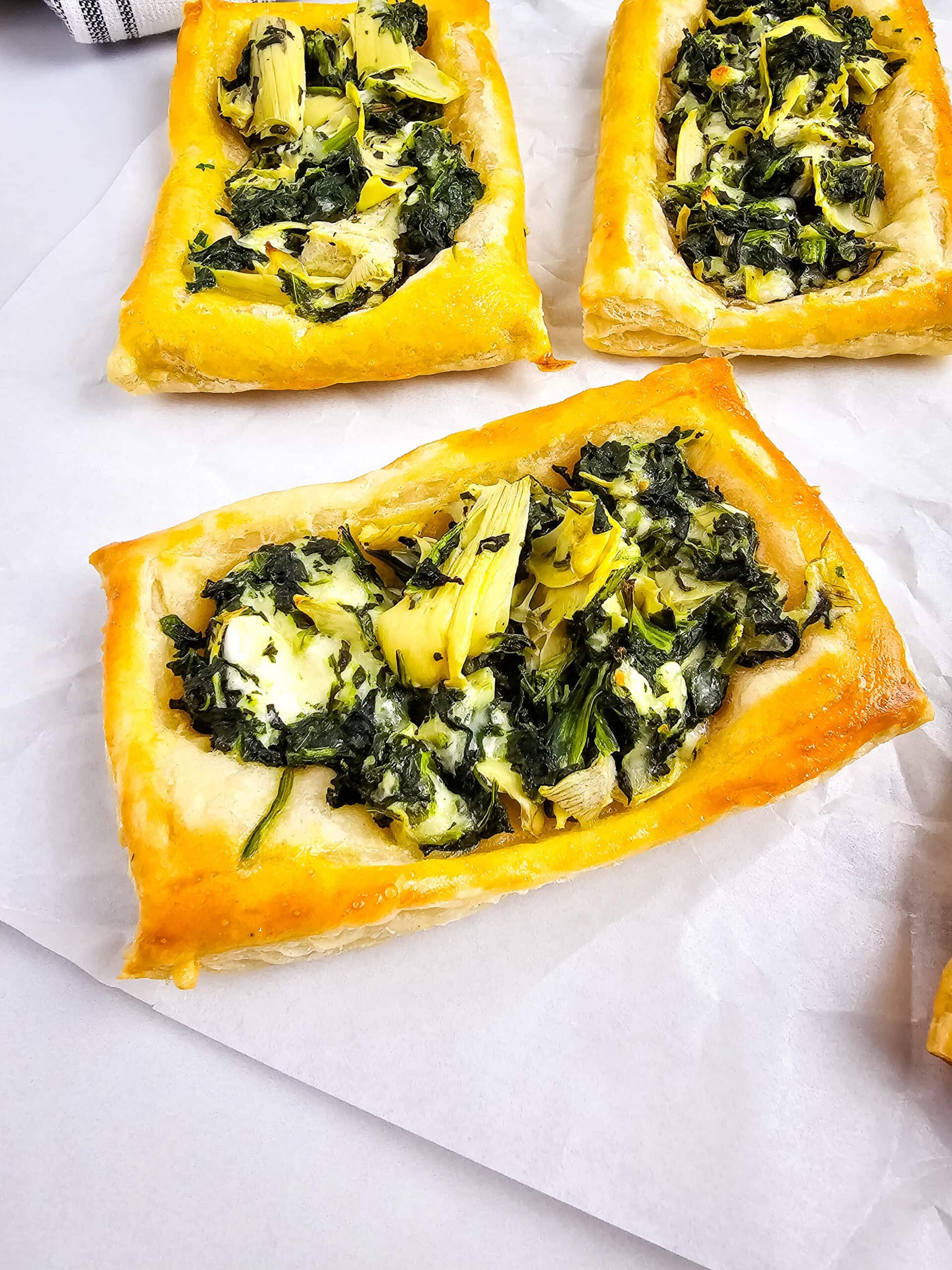 Three rectangular puff pastry tarts topped with a mixture of spinach, artichokes, and cheese, arranged on white parchment paper.
