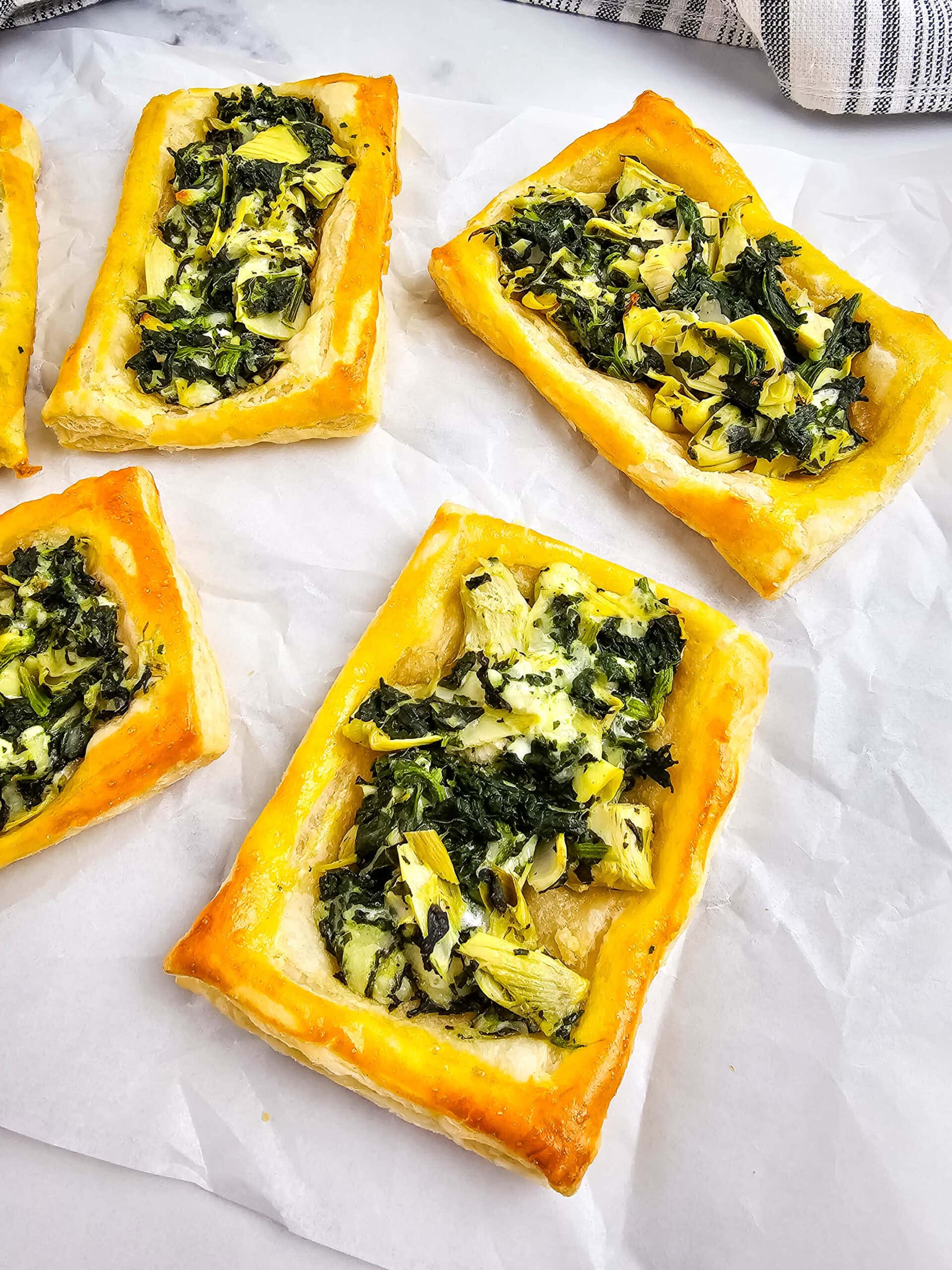 An overhead shot of rectangular puff pastry tarts topped with a mixture of spinach, artichoke, and melted cheese are arranged on white parchment paper. The pastries have golden brown, flaky edges.