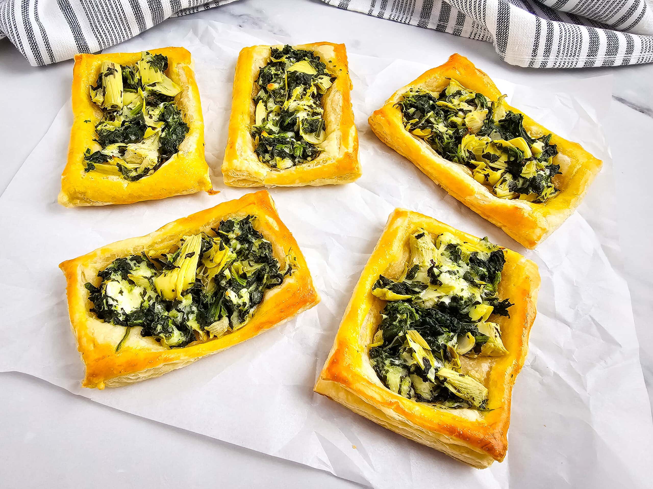 Five rectangular spinach and artichoke puff pastry tarts are arranged on a sheet of parchment paper, with a striped kitchen towel in the background. The tarts have golden, flaky edges and a savory filling.