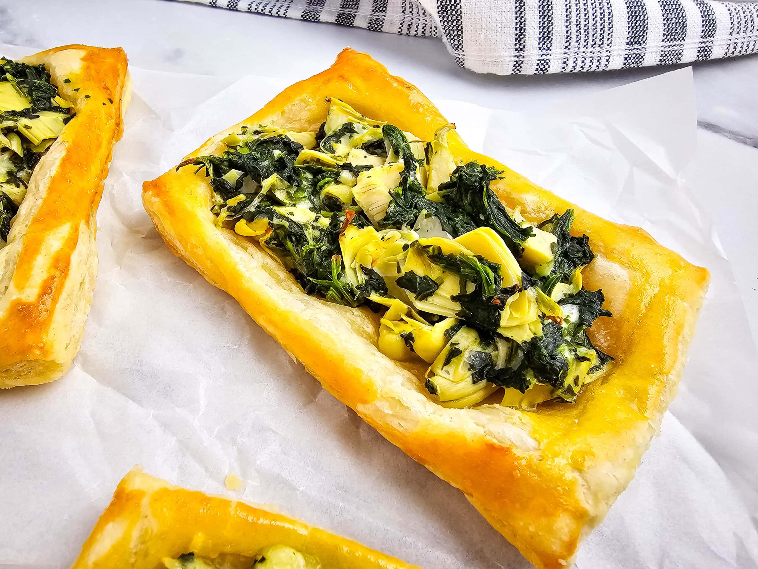 A rectangular puff pastry tart topped with spinach and artichoke filling sits on parchment paper, with a second tart partially visible nearby. A striped kitchen towel is in the background.
