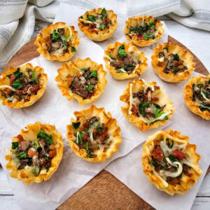 Mini phyllo pastry cups filled with sautéed mushrooms and spinach, bacon, and melted cheese are arranged on parchment paper atop a wooden board, with a striped cloth in the background.