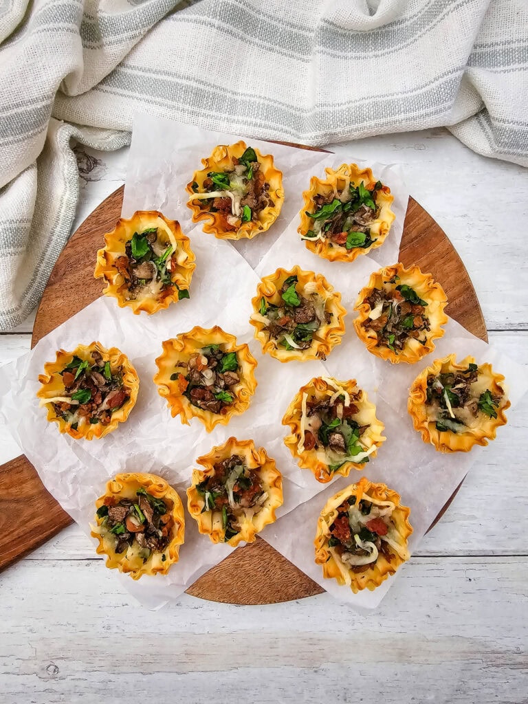 An overhead shot of twelve mini phyllo pastry cups filled with sautéed mushrooms and melted cheese are arranged on parchment paper atop a wooden board with a striped cloth in the background.
