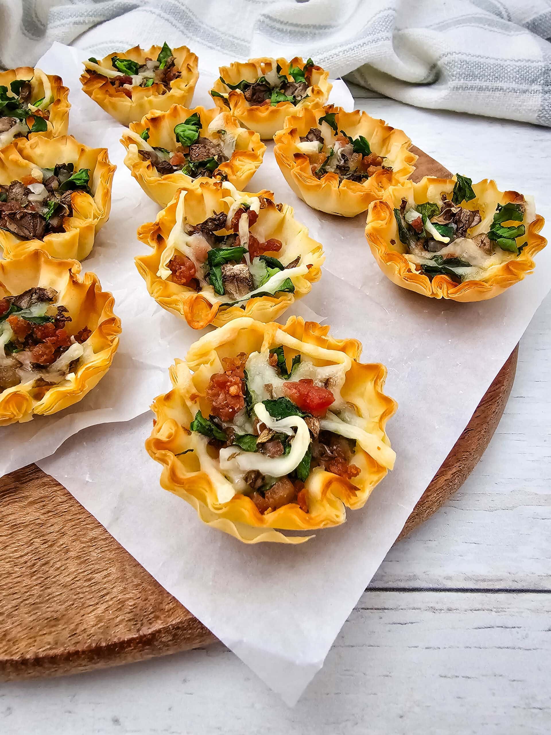 Mini phyllo pastry cups filled with melted cheese, chopped bacon, spinach, and mushrooms are arranged on white parchment paper atop a round wooden board, with a striped cloth in the background.