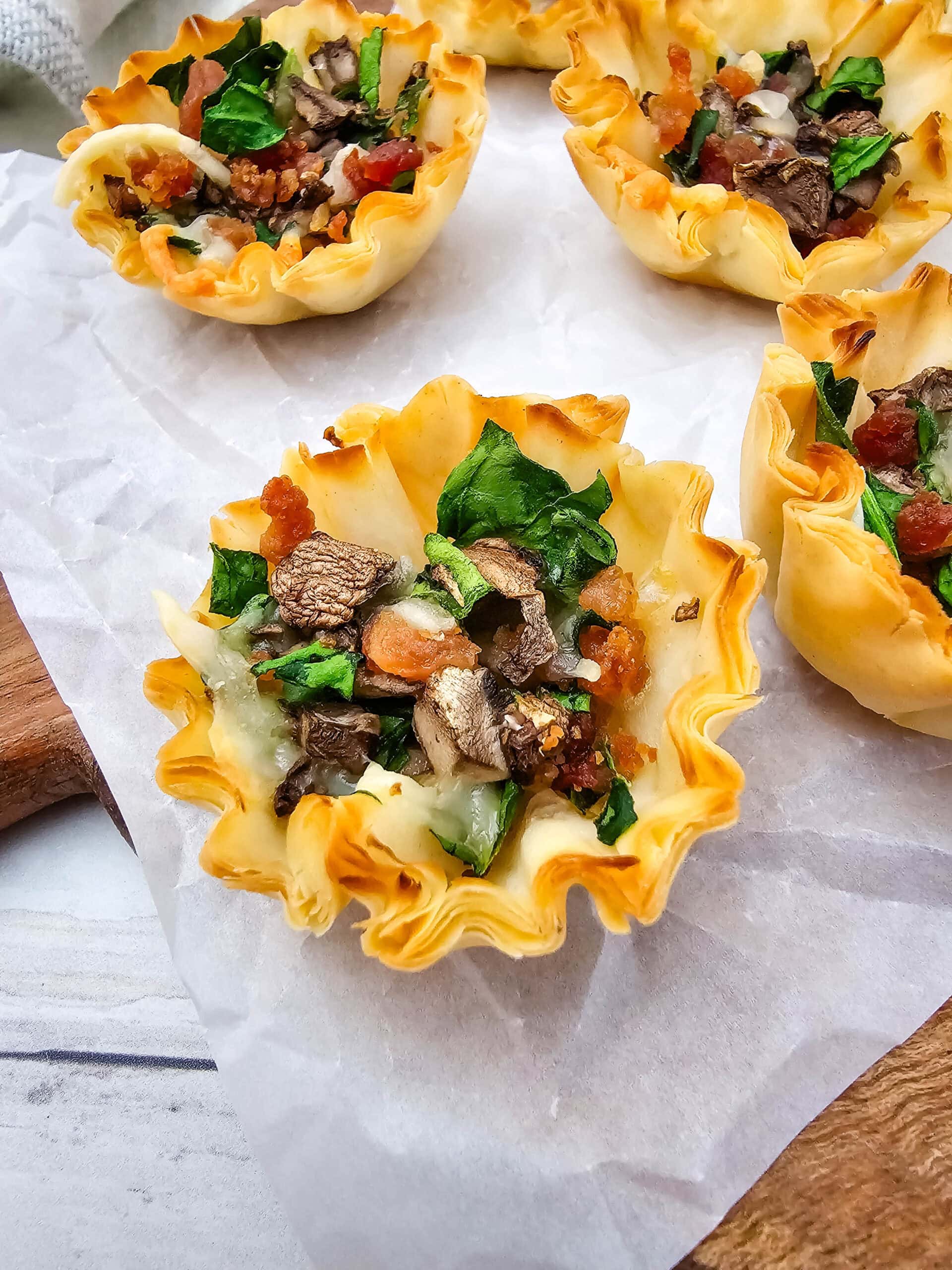 Phyllo pastry cups filled with chopped bacon, leafy greens, mushrooms, and melted cheese, arranged on parchment paper on a light wood surface.