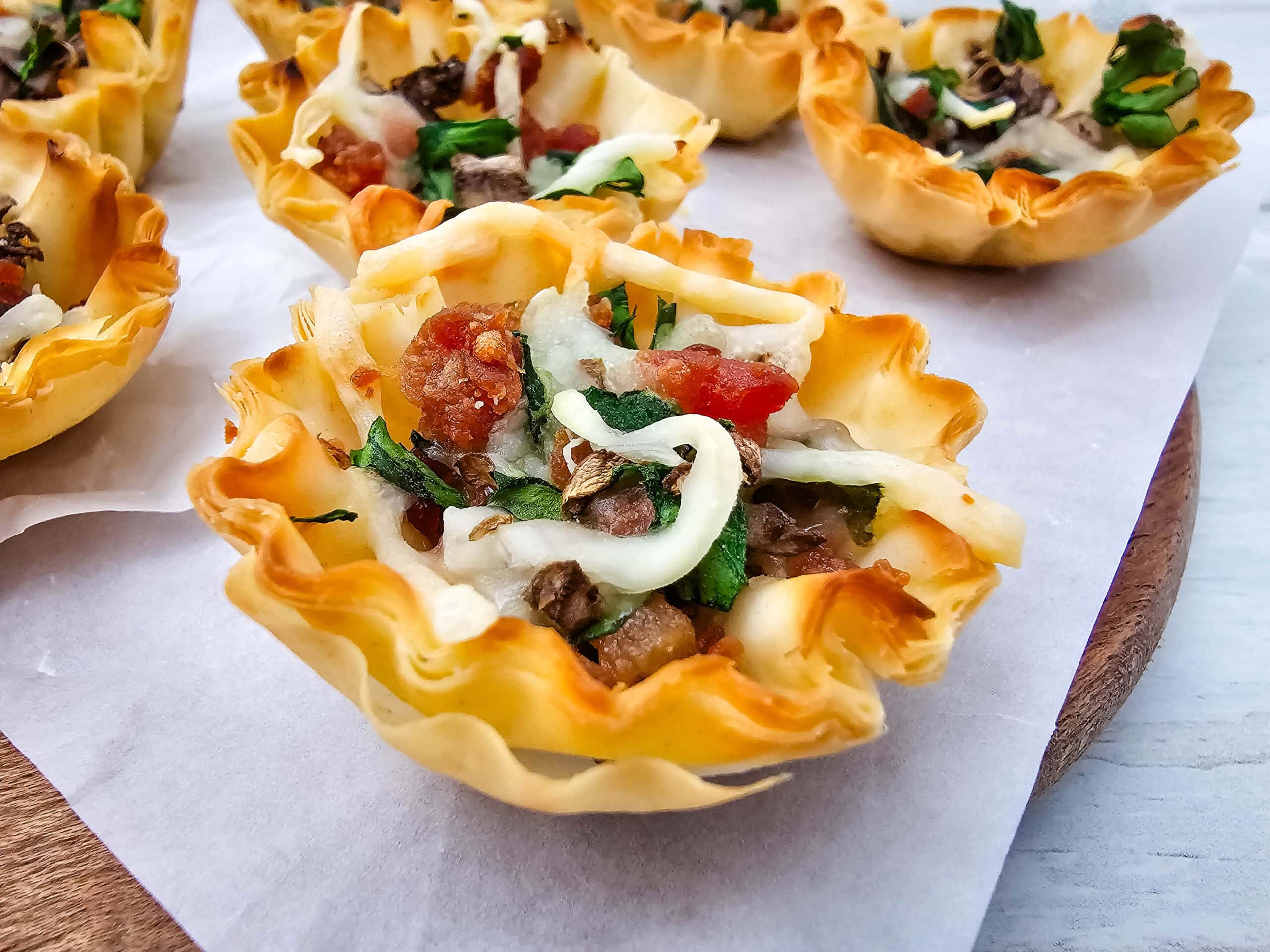 Close-up of crispy phyllo pastry cups filled with diced mushrooms and spinach, bacon, and melted cheese, arranged on a parchment-lined surface.