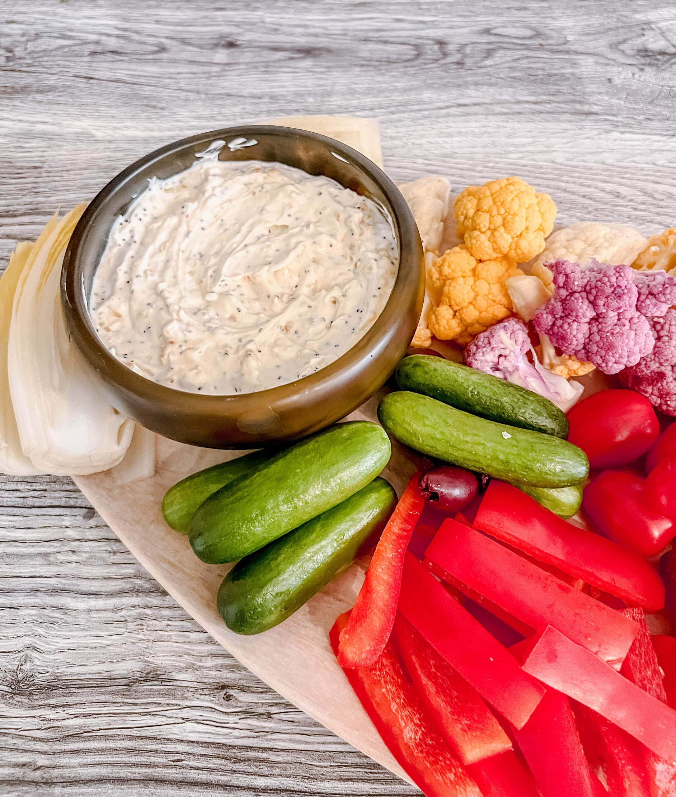 A platter with a bowl of creamy everything bagel dip surrounded by fresh vegetables, including mini cucumbers, red bell pepper strips, grape tomatoes, yellow and purple cauliflower, and endive on a wooden surface.