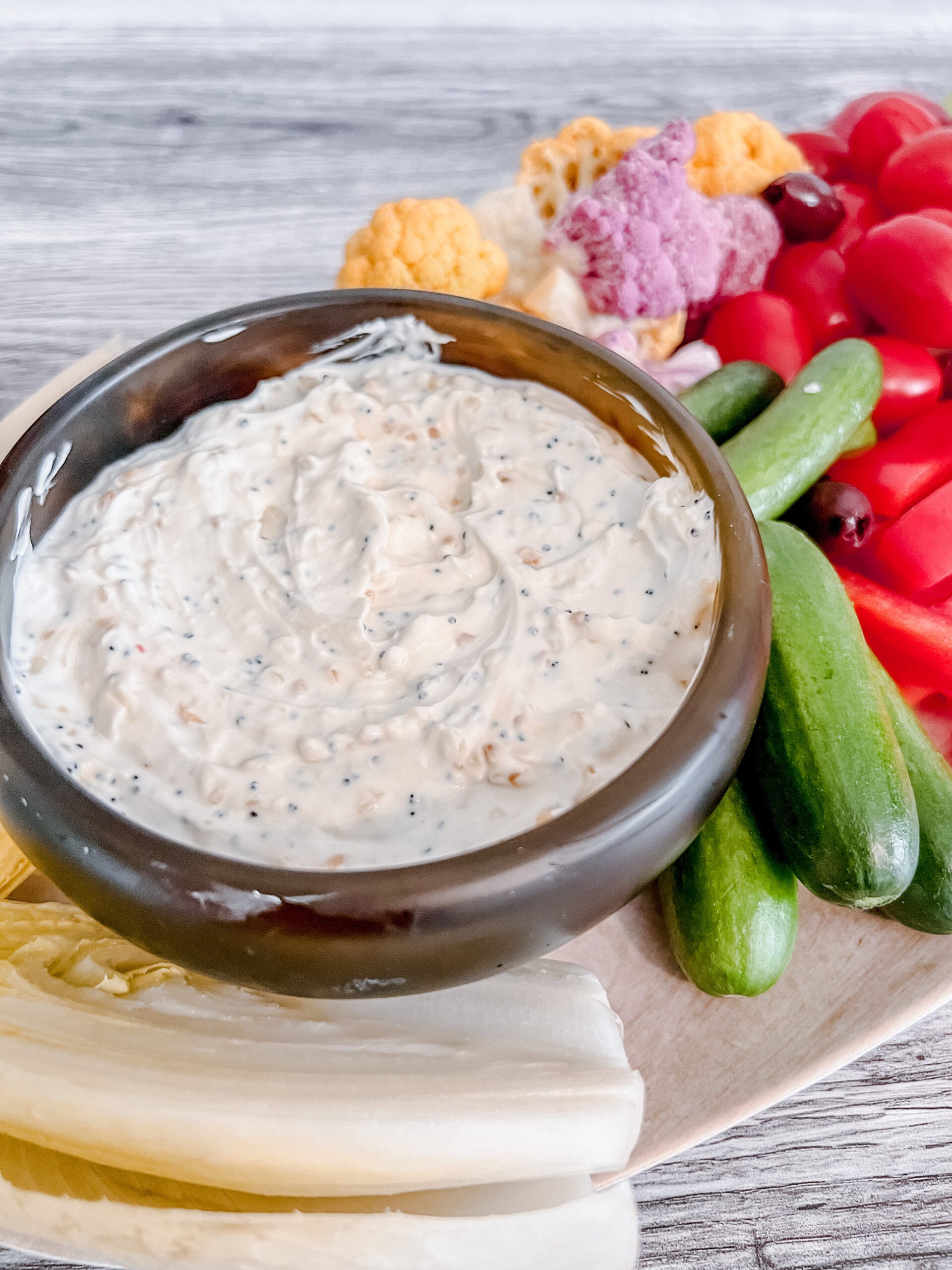 A bowl of creamy everything bagel dip sits on a platter surrounded by fresh vegetables, including mini cucumbers, cherry tomatoes, colorful cauliflower florets, and chicory, on a wooden surface.