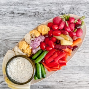 An oval wooden platter with colorful fresh vegetables&mdash;cauliflower, cherry tomatoes, radishes, mini cucumbers, red bell peppers, carrots, and olives&mdash;served with a bowl of creamy everything bagel dip, on a light wood surface.
