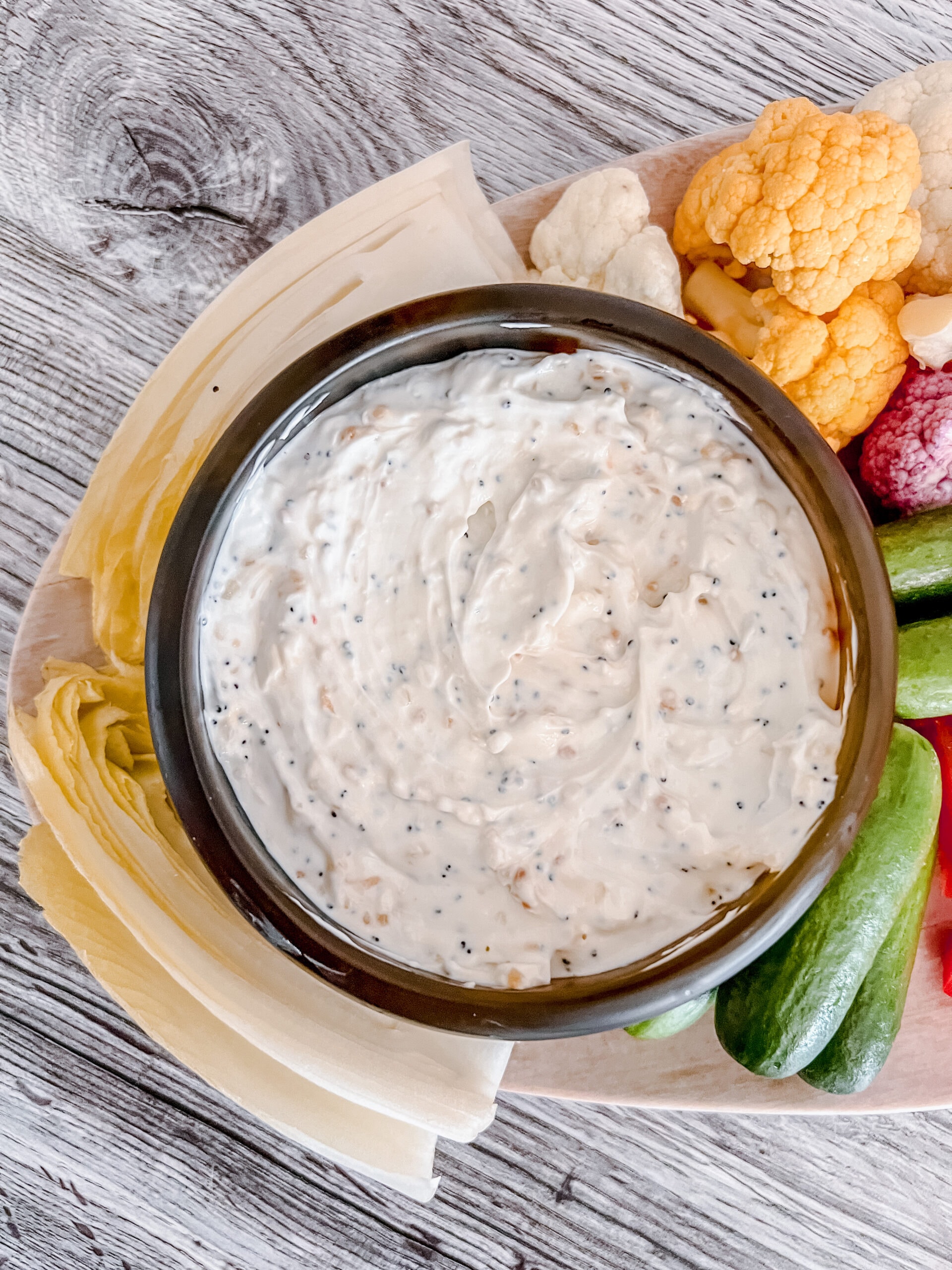 A bowl of creamy everything bagel dip sits on a wooden platter, surrounded by fresh vegetables including endive, cucumbers, cauliflower, and carrots, ready for dipping.
