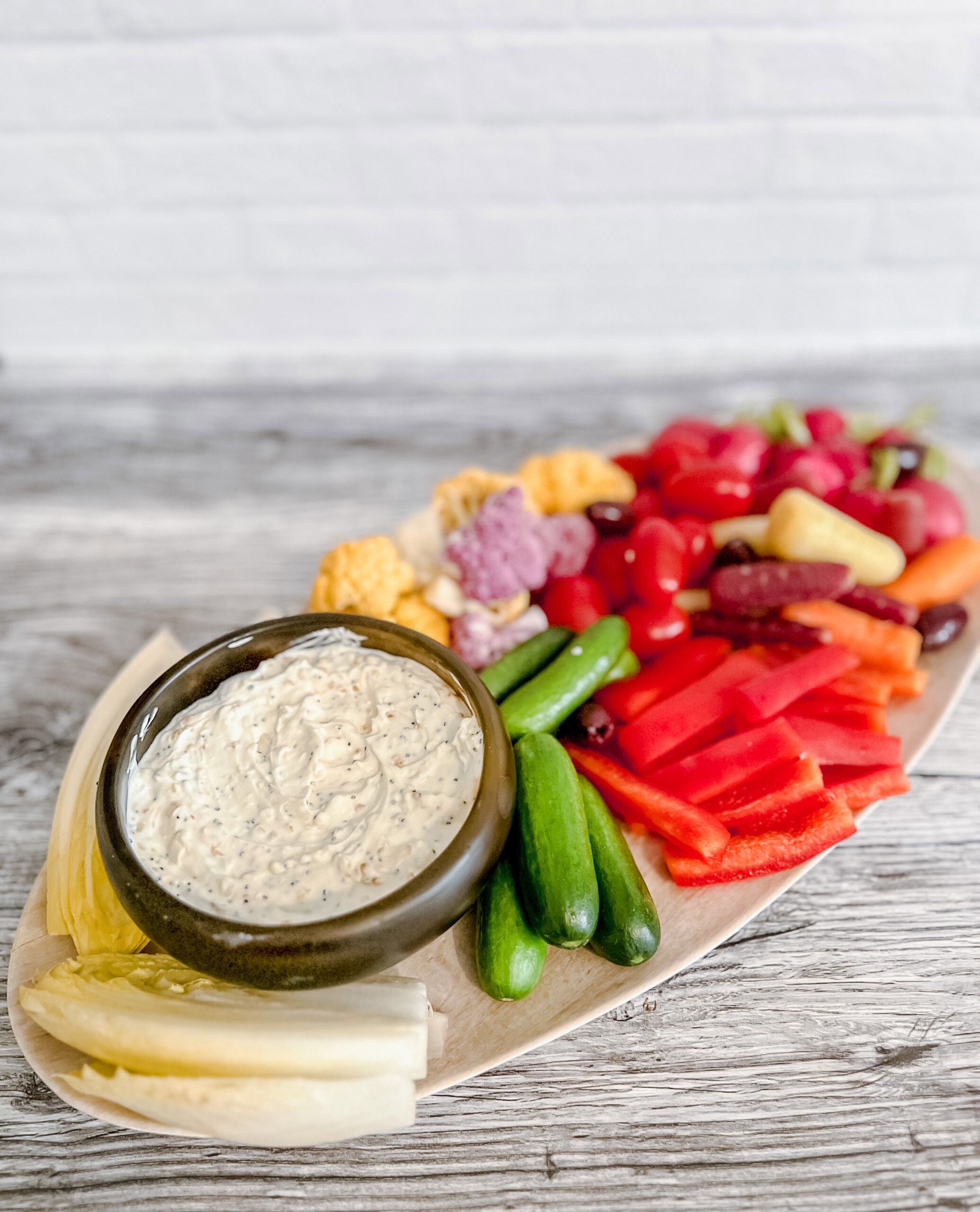 A wooden platter with assorted fresh vegetables&mdash;cucumbers, red bell peppers, cherry tomatoes, carrots, cauliflower, and yellow peppers&mdash;next to a bowl of creamy everything bagel dip. The background is a light wooden surface.