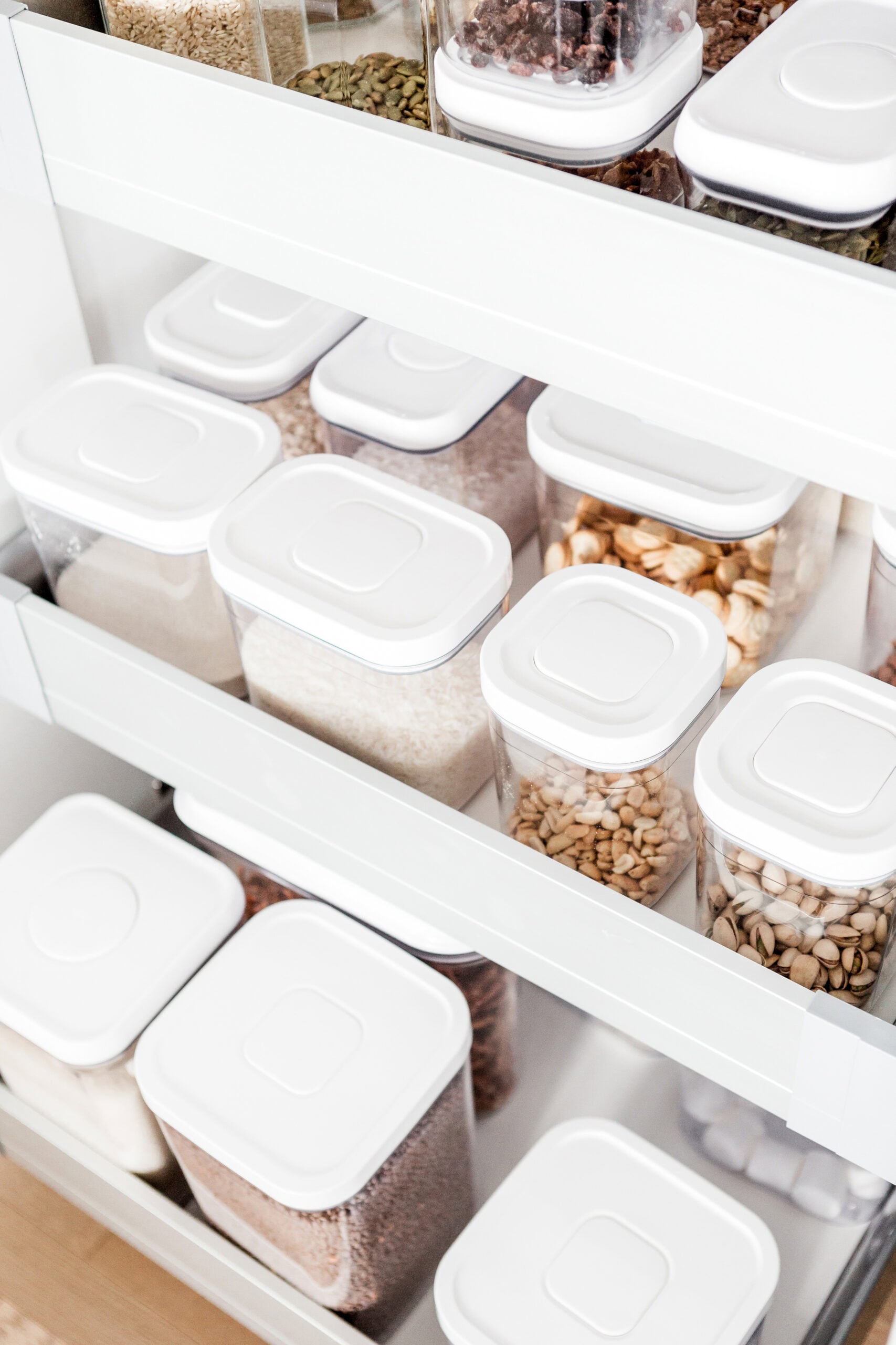 Clear plastic containers with white lids are neatly organized on white pantry shelves, holding various dry goods such as grains, seeds, nuts, and beans.