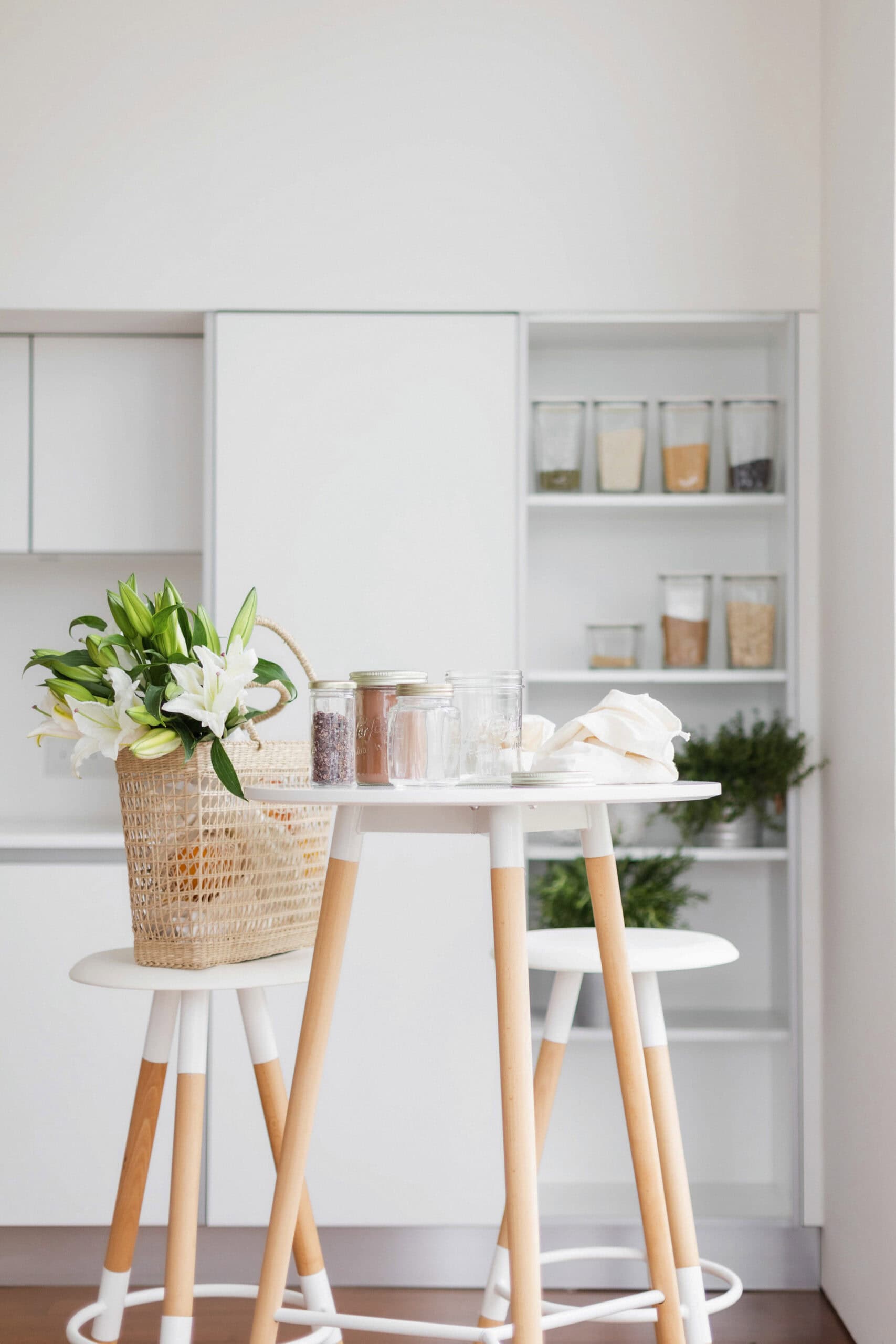 A bright, minimalist kitchen features a small table with wooden stools, a basket of white lilies, glass jars, and a napkin. A table between the two stools holds some glass jars and a white cloth. Shelves in the background hold jars with grains and herbs. The overall color scheme is white and light wood.