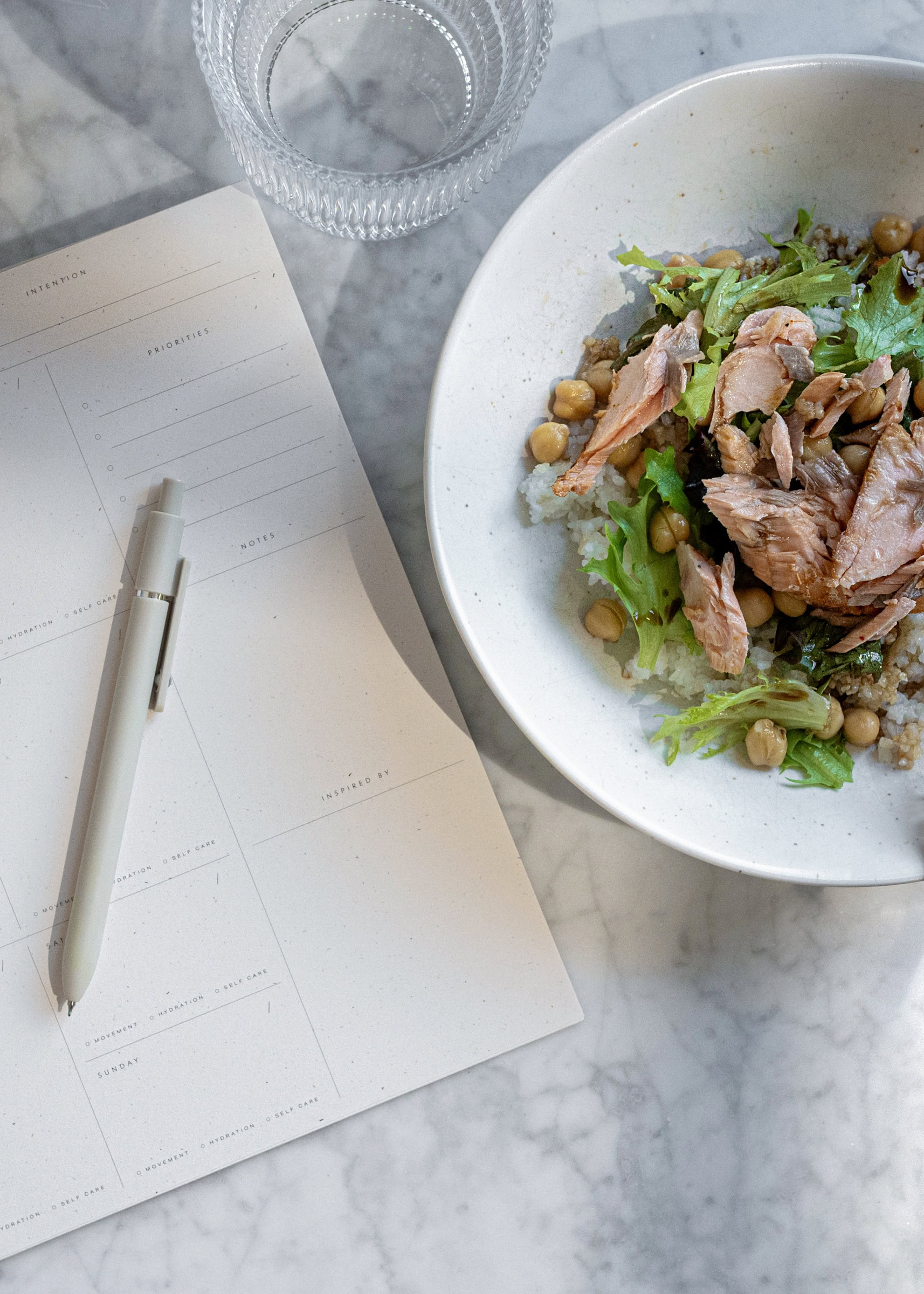 A bowl of salad with greens, chickpeas, and shredded meat sits on a marble surface next to a planning notepad with a pen and water glass.