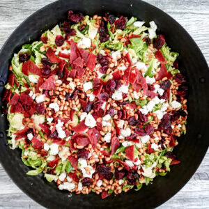A large black bowl filled with a colorful salad of shredded greens, crumbled cheese, dried cranberries, pine nuts, and pieces of turkey bacon, set on a gray wooden surface.