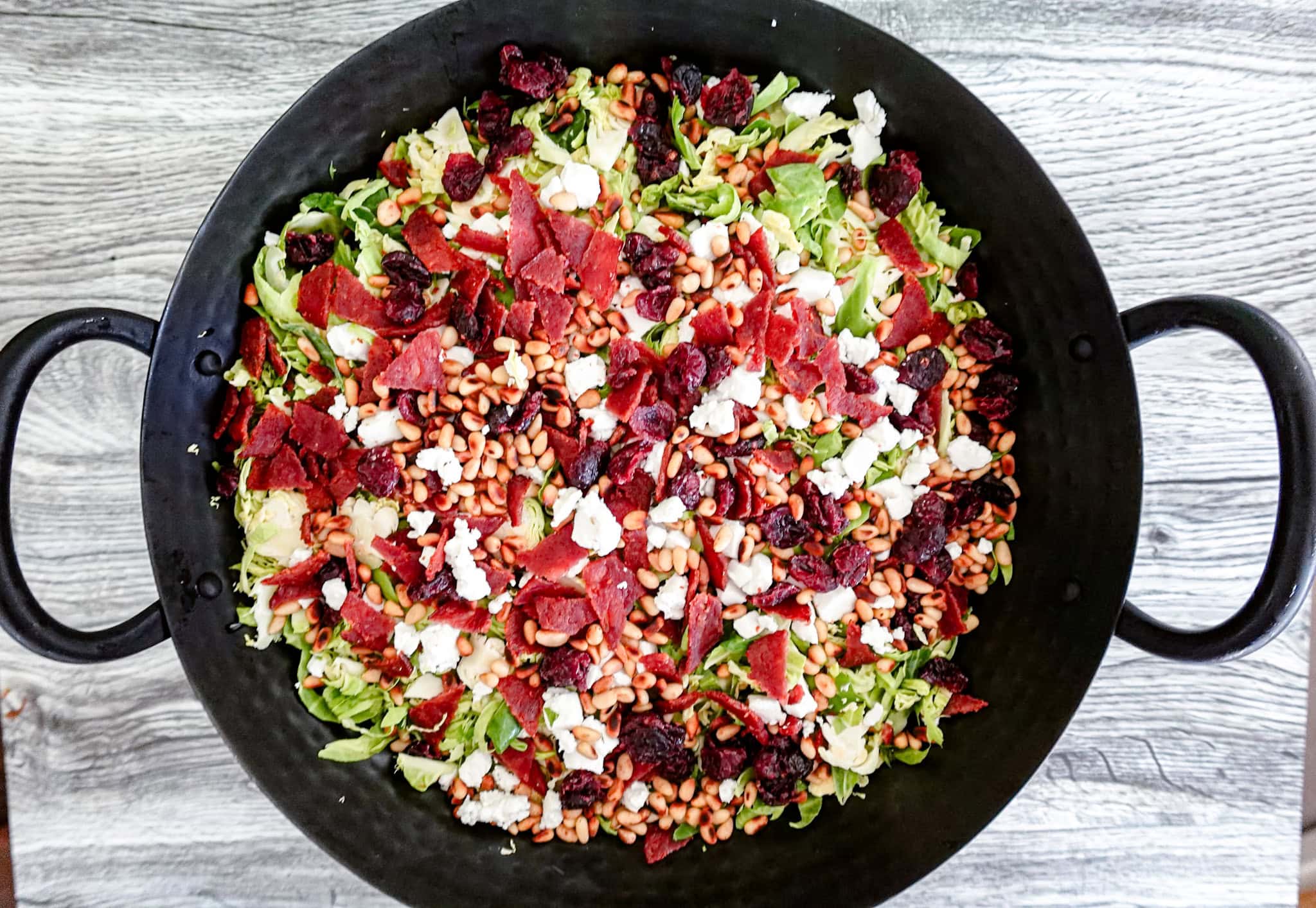 A large black bowl filled with a colorful salad of shredded greens, crumbled cheese, dried cranberries, pine nuts, and pieces of turkey bacon, set on a gray wooden surface.