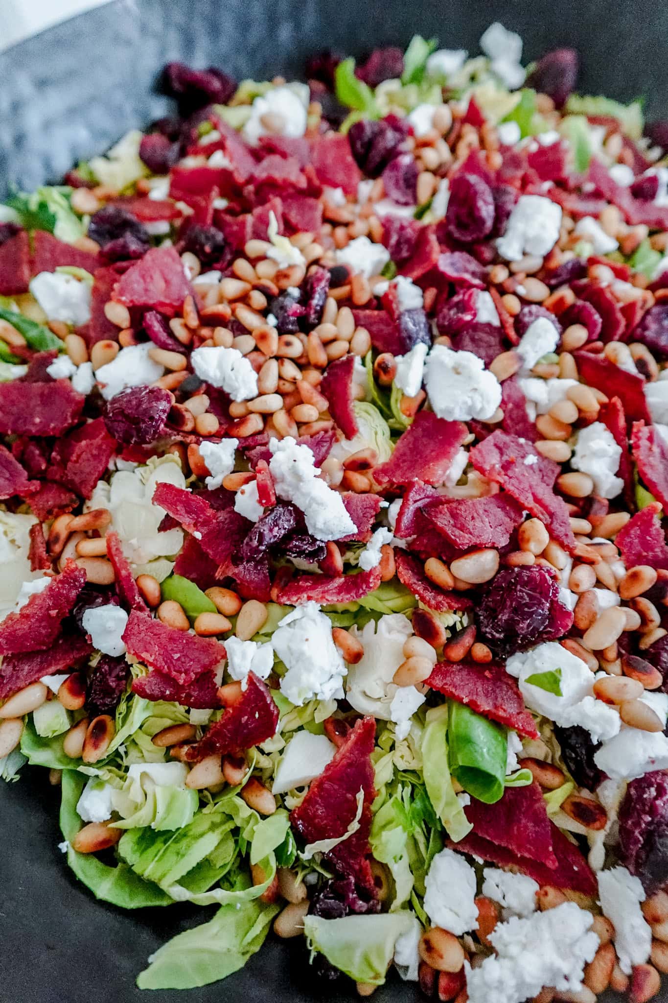A close-up of a salad topped with pine nuts, dried cranberries, crumbled feta cheese, and pieces of dried or cured meat on a bed of sliced Brussels sprouts.