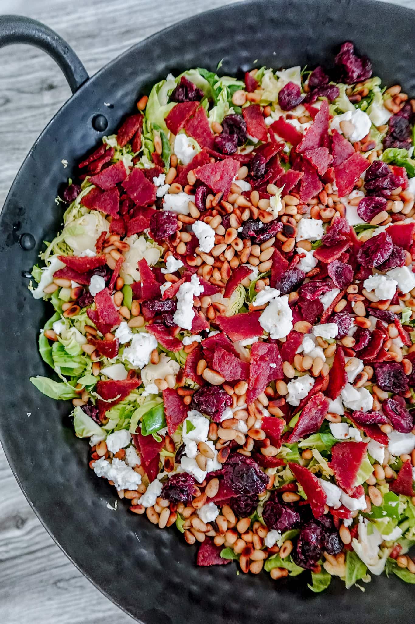 A close-up of a salad in a black bowl, featuring shredded Brussels sprouts, pine nuts, crumbled white cheese, dried cranberries, and pieces of crispy red bacon. The bowl sits on a gray wooden surface.