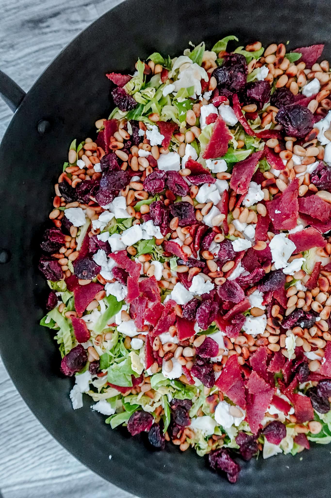 A close-up of a salad in a black bowl, featuring shredded Brussels sprouts, goat cheese, pine nuts, dried cranberries, and pieces of cooked bacon. The ingredients are mixed and colorful.