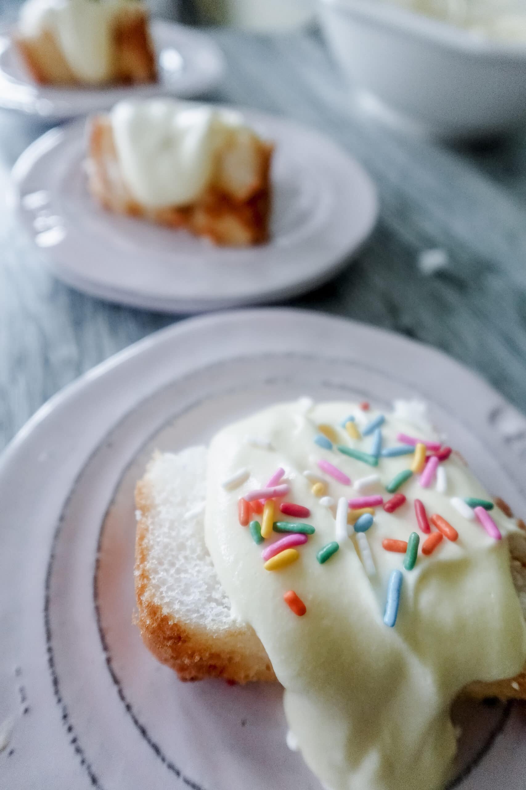 A close-up of a slice of cake topped with Bavarian Cream and colorful sprinkles on a plate, with two more similar slices on plates in the blurred background.