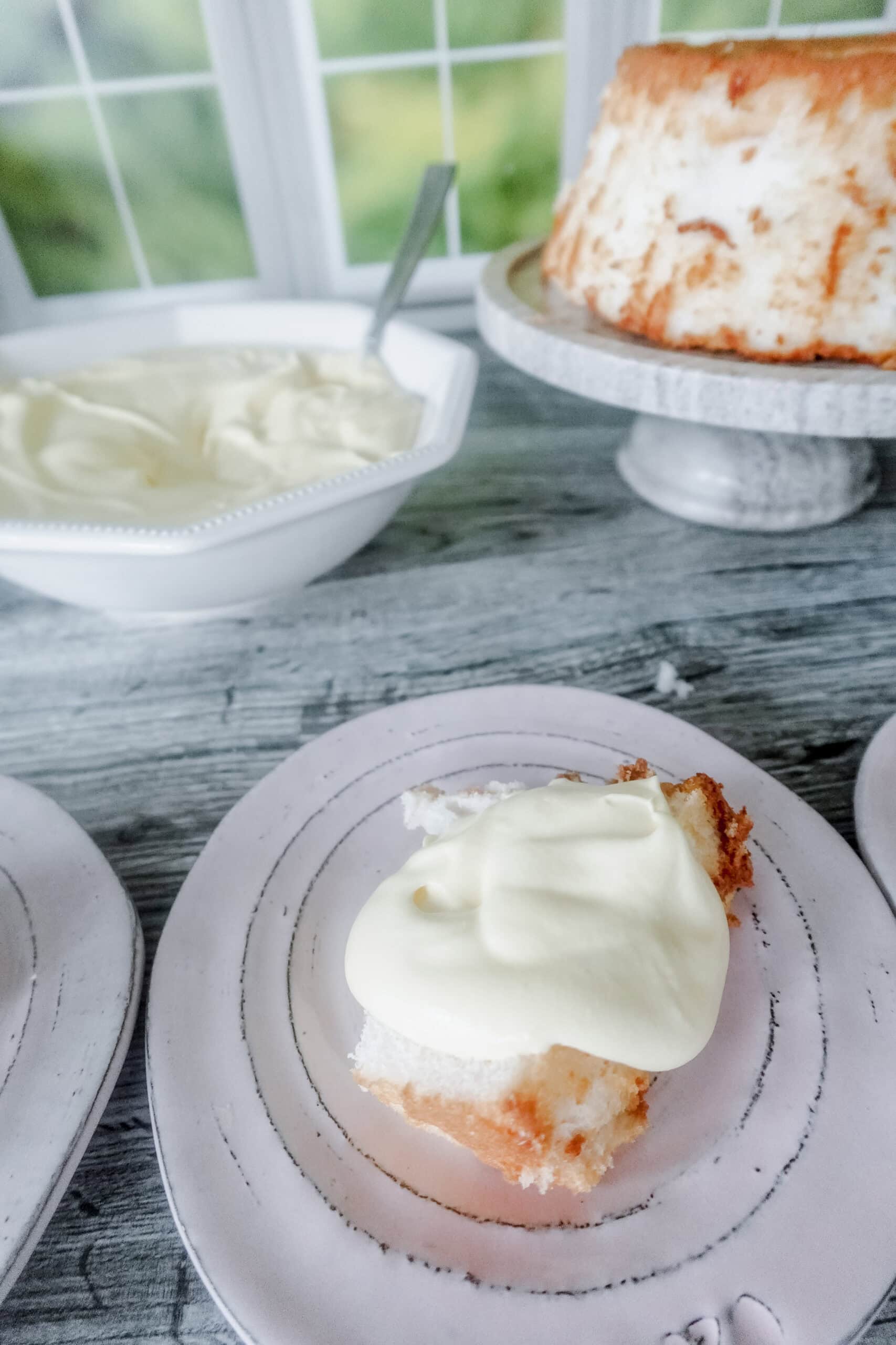 A slice of angel food cake topped with Bavarian cream sits on a plate, with a bowl of more cream and the rest of the cake on a stand in the background, near a window.
