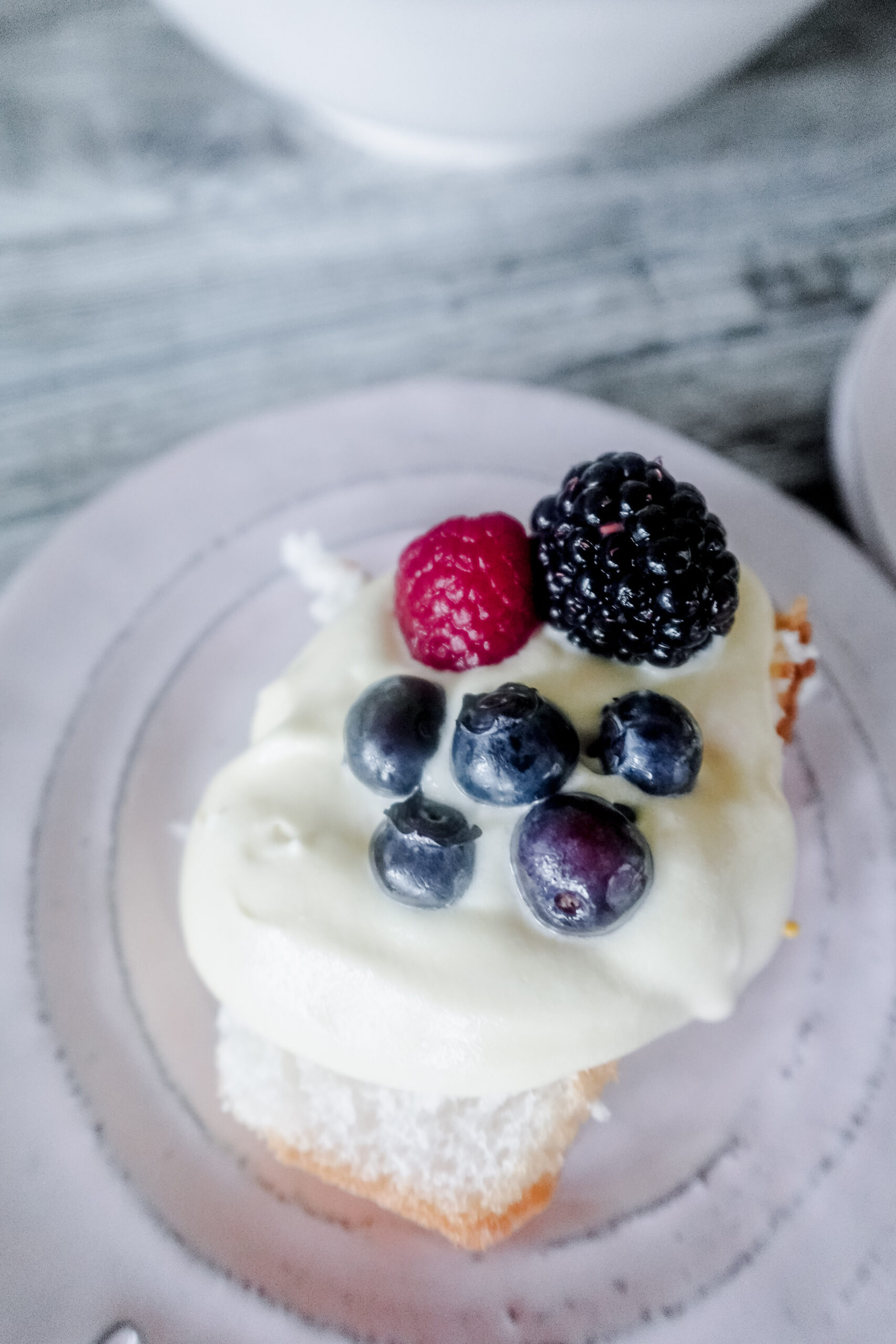 A slice of cake topped with Bavarian cream, blueberries, a raspberry, and a blackberry, served on a light-colored plate.