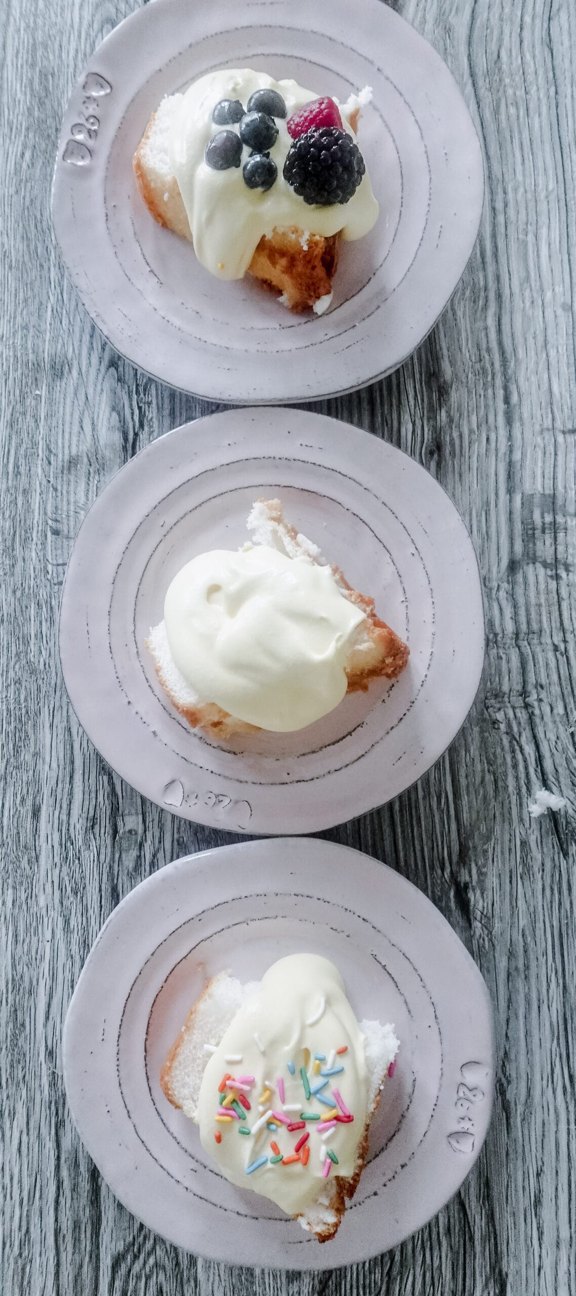 Overhead shot of three small plates with slices of cake topped with Bavarian cream: the top slice has mixed berries, the middle has plain cream, and the bottom has colorful sprinkles. All plates are on a rustic wooden surface.
