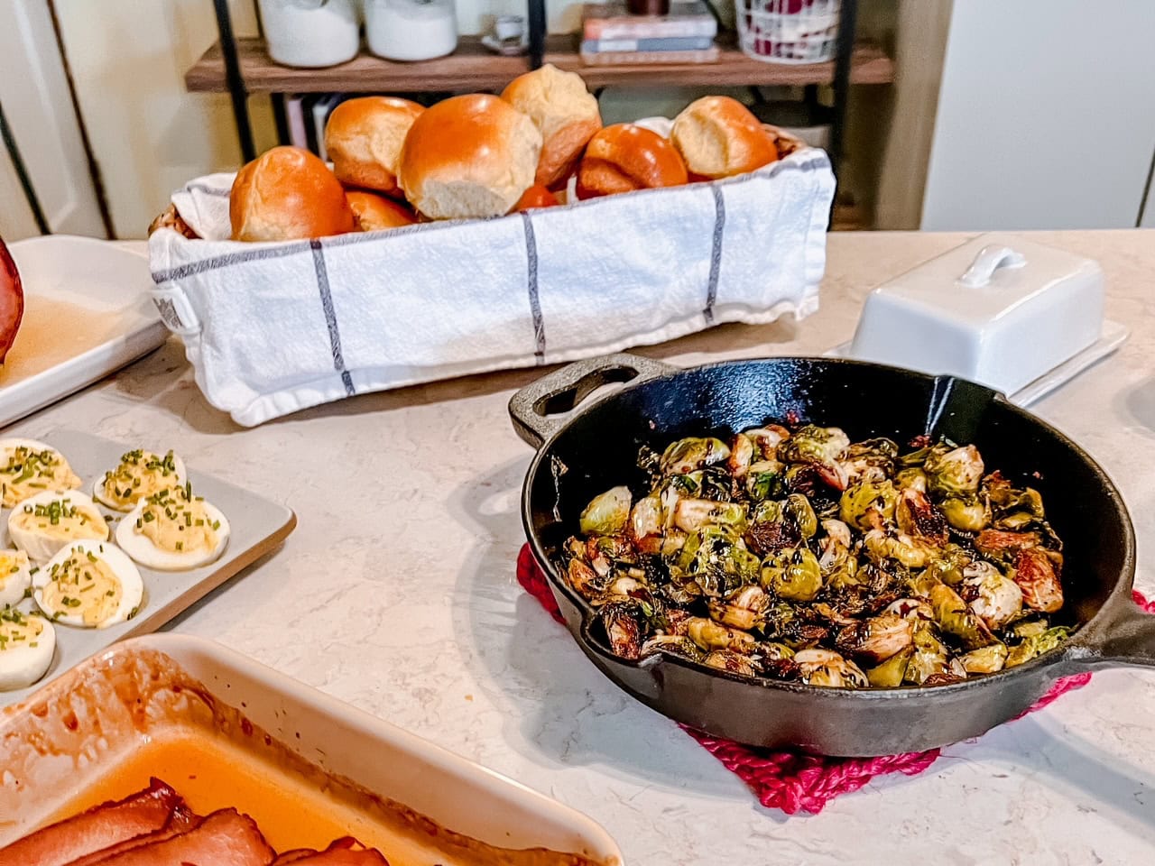 A cast-iron skillet with roasted Brussels sprouts sits on a countertop next to a basket of bread rolls, a dish of deviled eggs, a covered butter dish, and a tray of sliced ham.