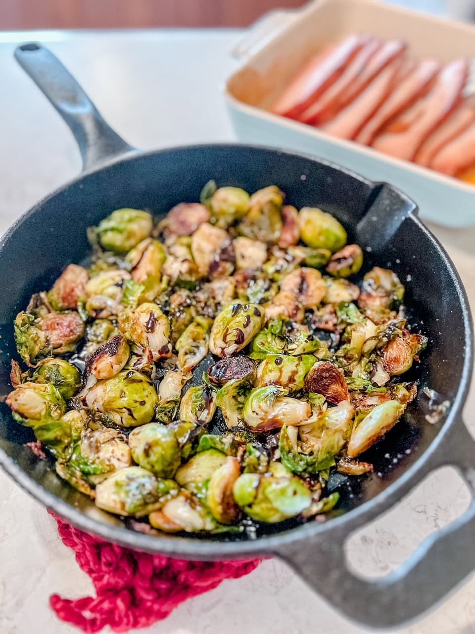 Brussels sprouts saut&eacute;ed in a black cast-iron skillet, garnished and lightly browned, sit on a red trivet. In the background, a dish with thick-sliced ham strips lay in a baking pan.