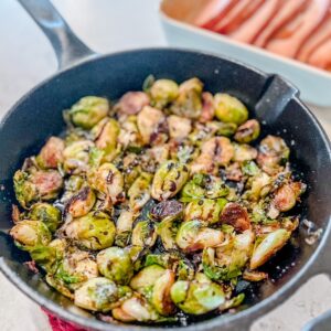 A cast-iron skillet filled with roasted Brussels sprouts sits on a red mat. In the background, thick-sliced ham are visible in a white dish.