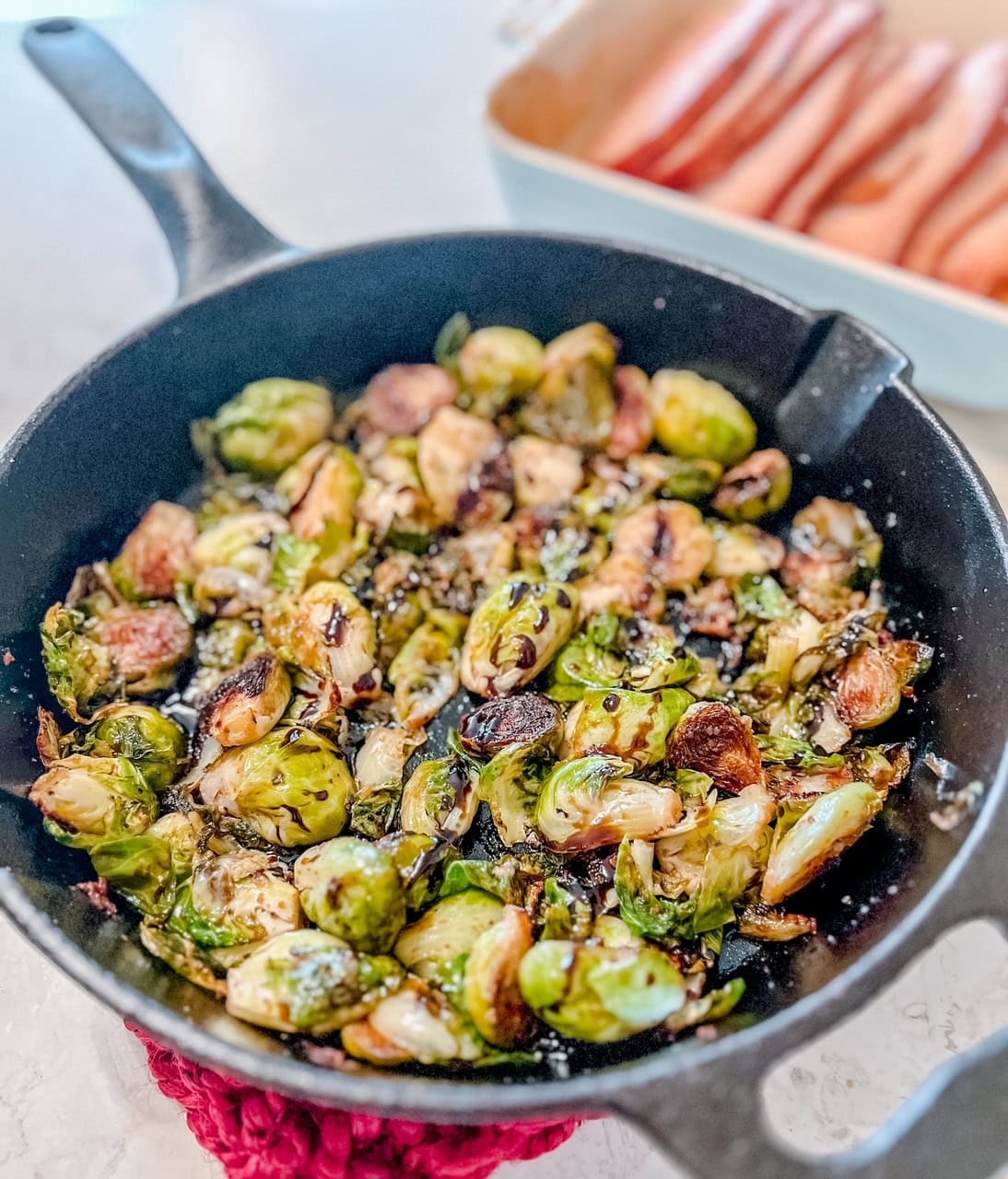 A cast-iron skillet filled with roasted Brussels sprouts sits on a red mat. In the background, thick-sliced ham are visible in a white dish.