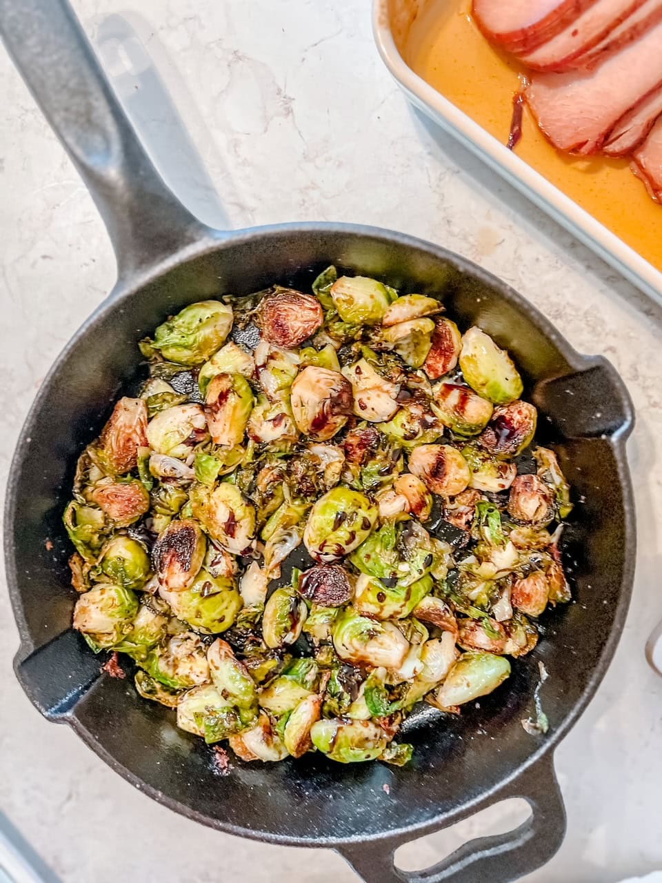 Overhead shot of a cast-iron skillet filled with roasted Brussels sprouts, some charred and crispy, sits on a light countertop next to a dish of sliced ham.