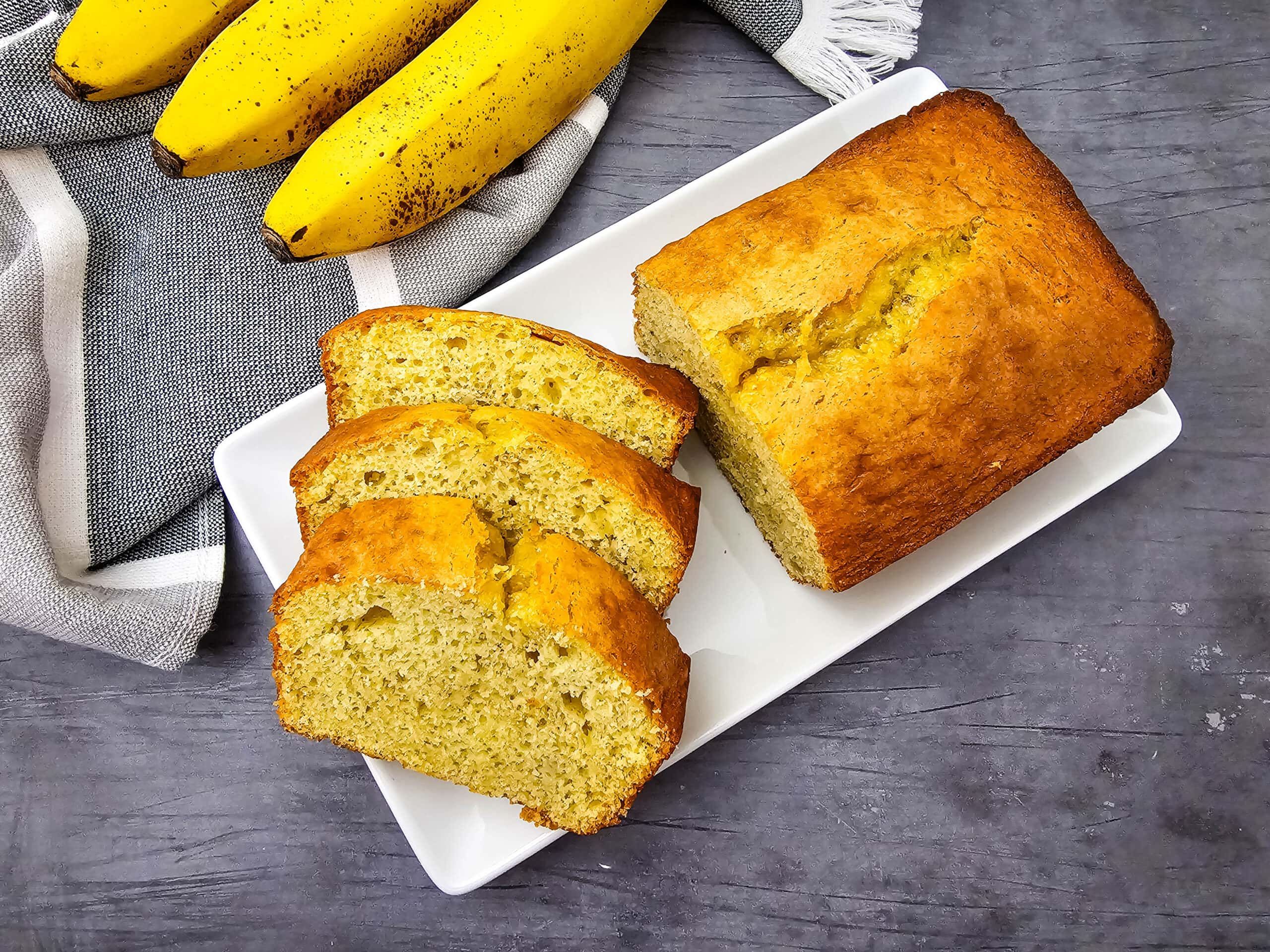 Overhead shot of a loaf of banana bread with three slices cut, served on a white plate. Whole bananas and a gray and white striped towel are in the background, set on a textured gray surface.