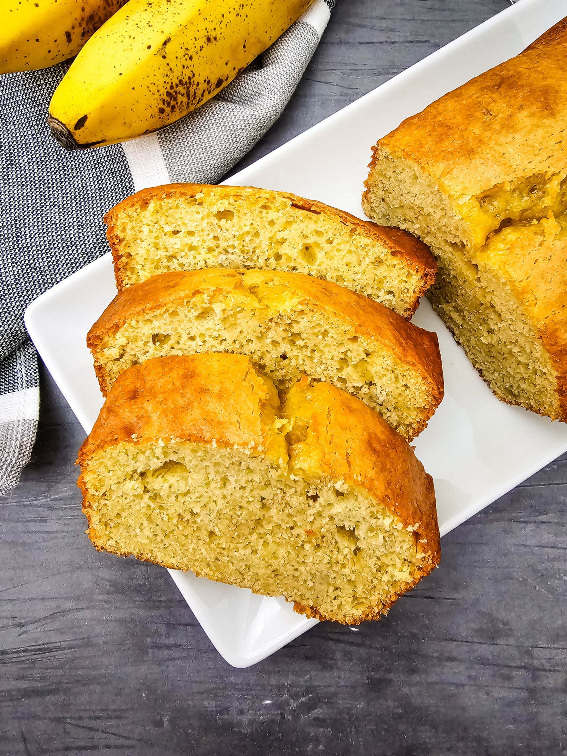 An overhead shot of three slices of banana bread that are arranged on a white plate next to a loaf, with ripe bananas and a striped cloth in the background, all on a gray surface.