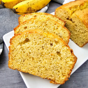Three slices of banana bread are arranged on a white plate, with the rest of the loaf behind them. In the background, ripe bananas and a striped kitchen towel are visible on a gray surface.