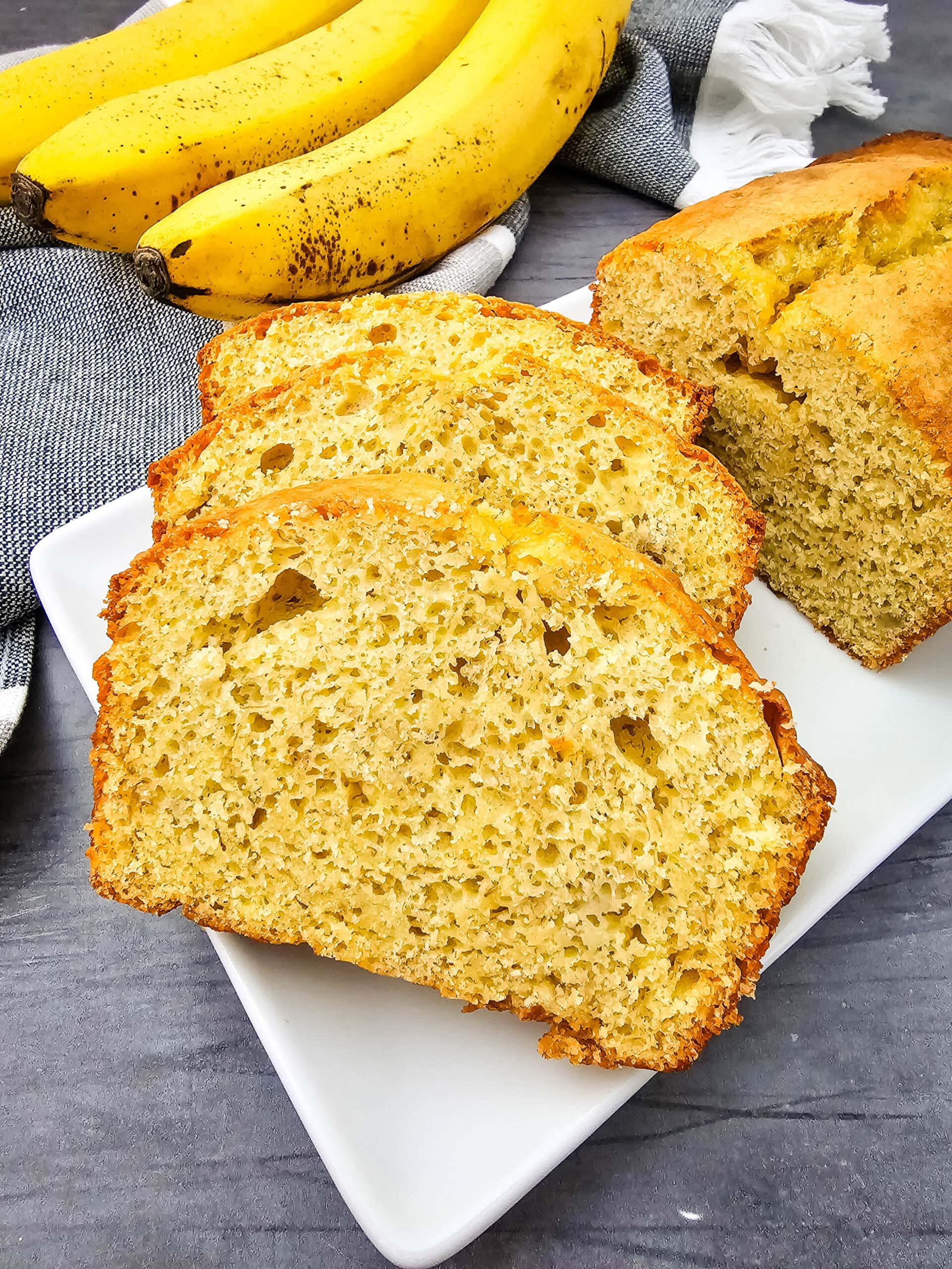 Three slices of banana bread are arranged on a white plate, with the rest of the loaf behind them. In the background, ripe bananas and a striped kitchen towel are visible on a gray surface.