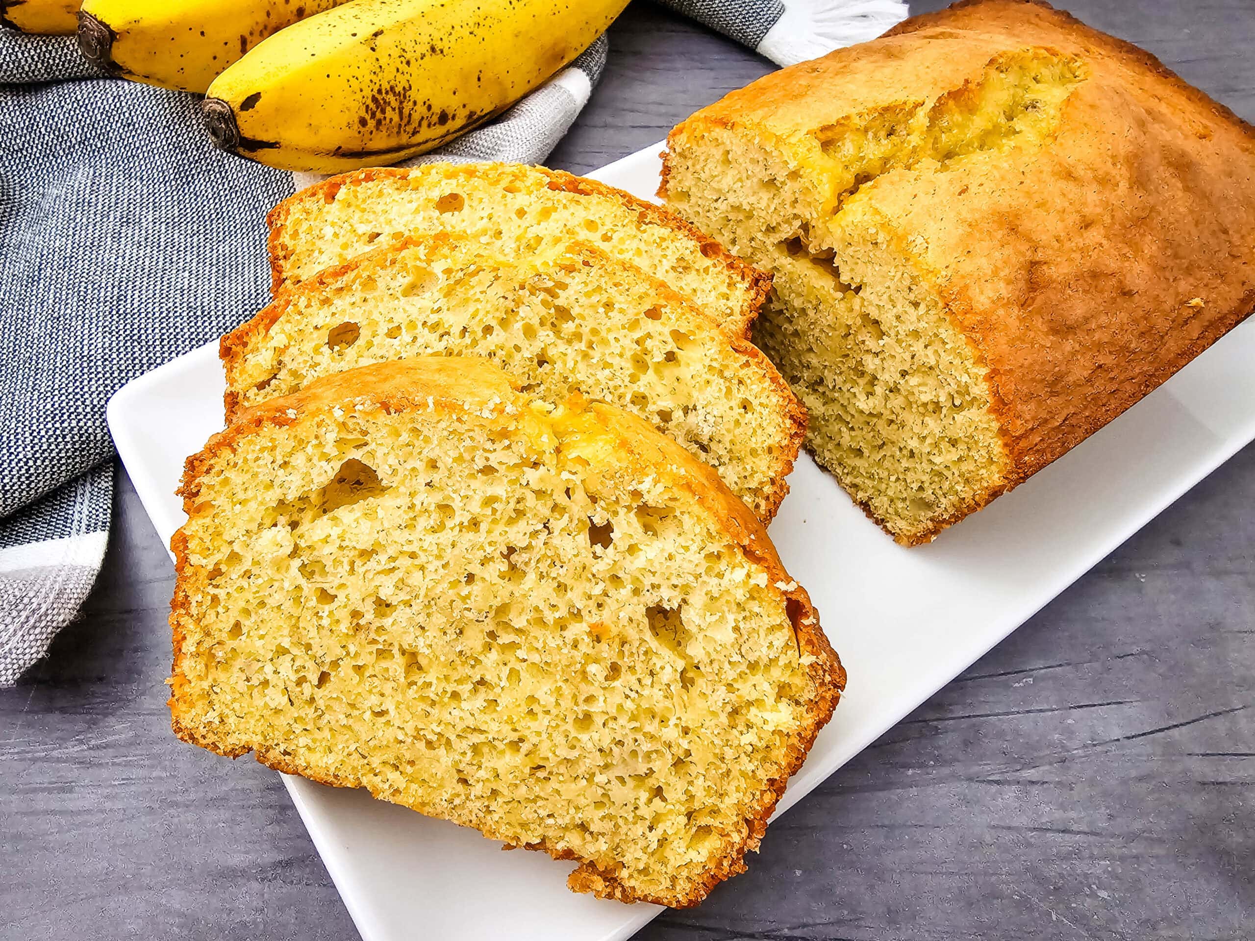 A loaf of banana bread sits on a white plate, with three slices cut and fanned out in front. Ripe bananas and a gray towel are in the background on a gray surface.