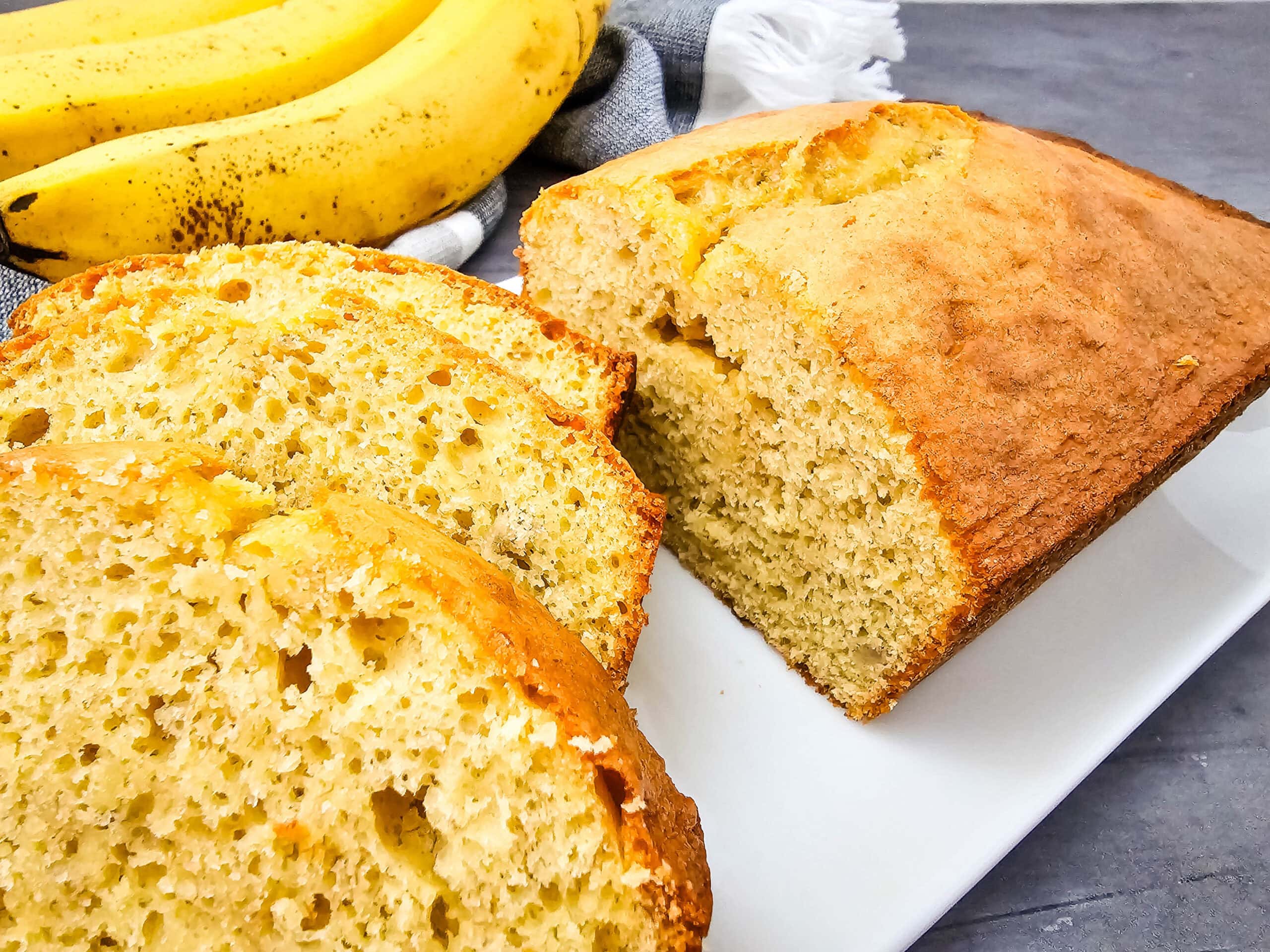 A close-up of a loaf of banana bread, partially sliced, is displayed on a white plate. Three whole ripe bananas and a gray towel are in the background on a gray surface.