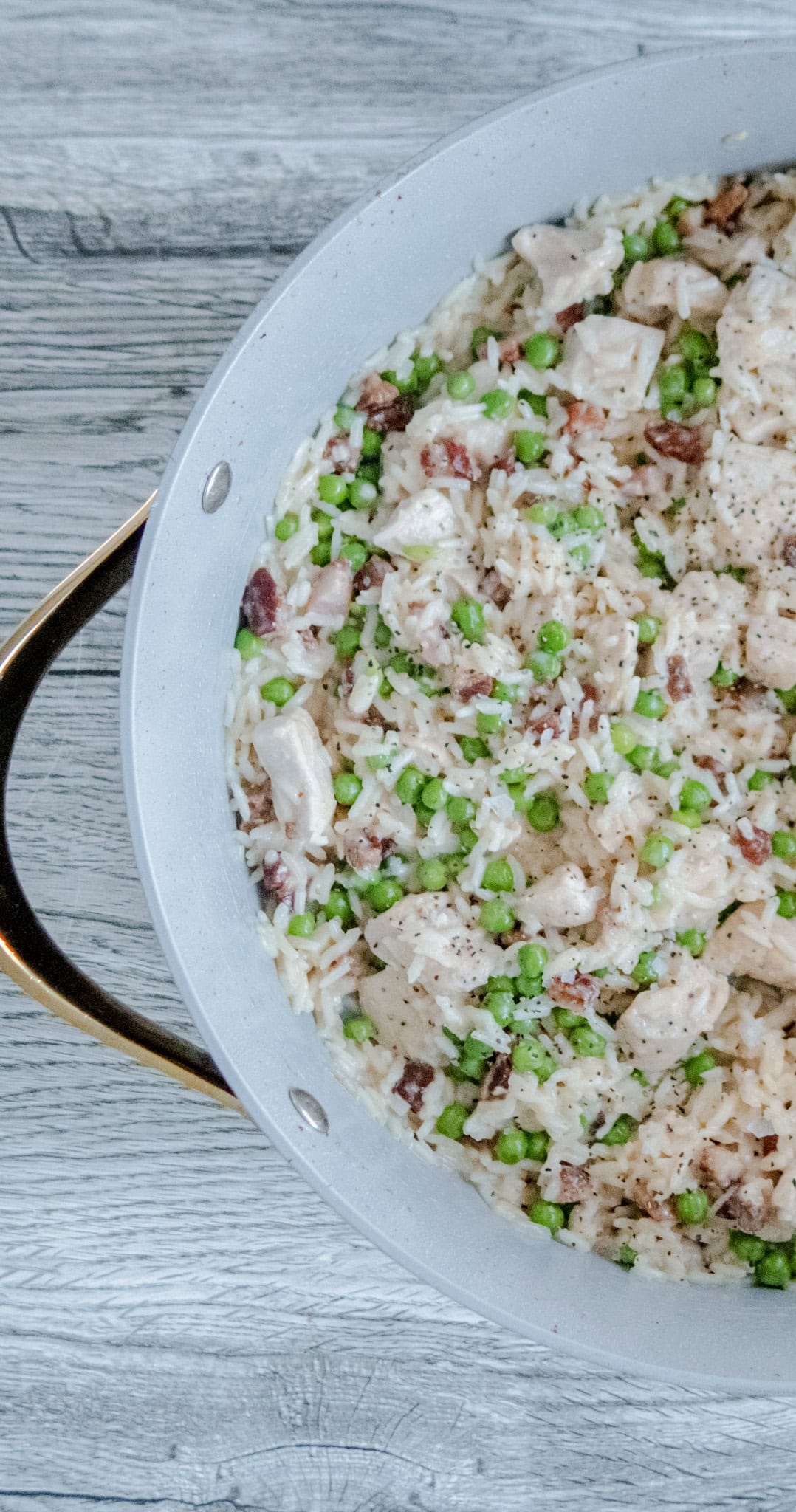 An overhead shot of half a pan filled with creamy rice, green peas, bacon bits, and chunks of chicken, sitting on a light wood surface.