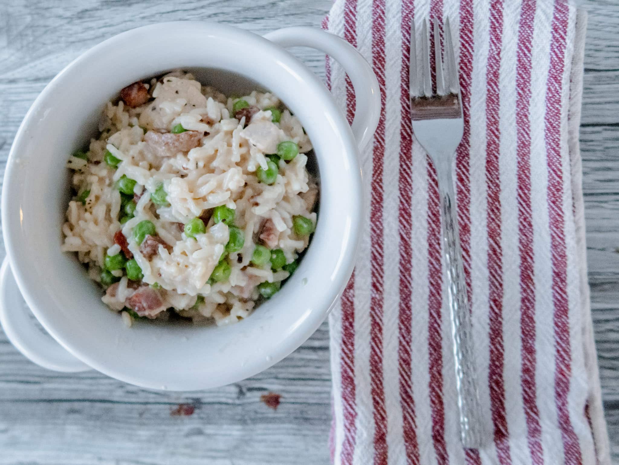 A white bowl filled with rice, peas, and pieces of chicken and crispy pancetta sits on a light wooden surface next to a metal fork placed on a red-and-white striped cloth napkin.