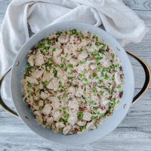 An overhead shot of a large, white pan filled with creamy rice, diced chicken, green peas, and bits of pancetta, set on a light wooden surface with a white cloth beside it.