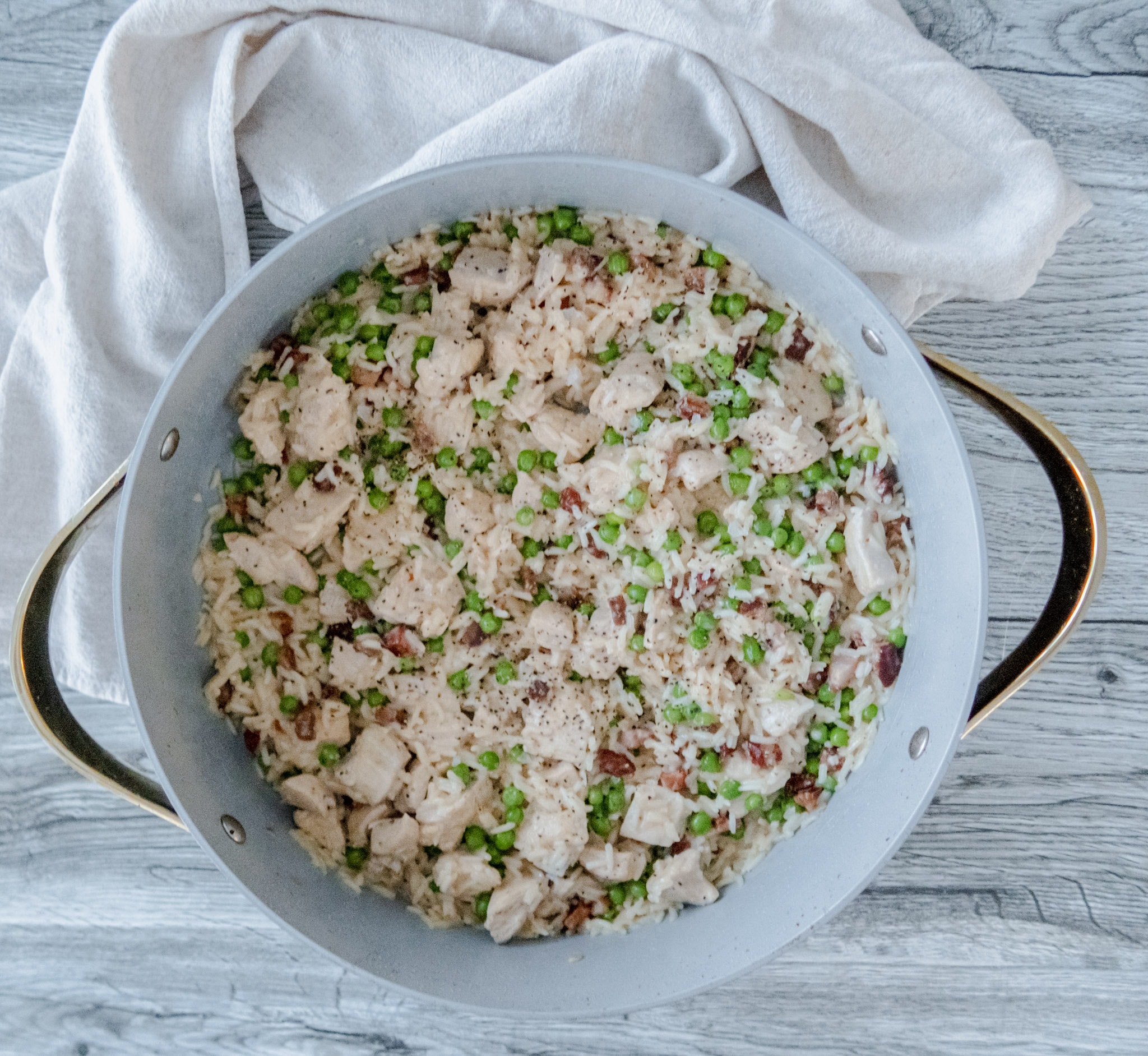 An overhead shot of a large, white pan filled with creamy rice, diced chicken, green peas, and bits of pancetta, set on a light wooden surface with a white cloth beside it.