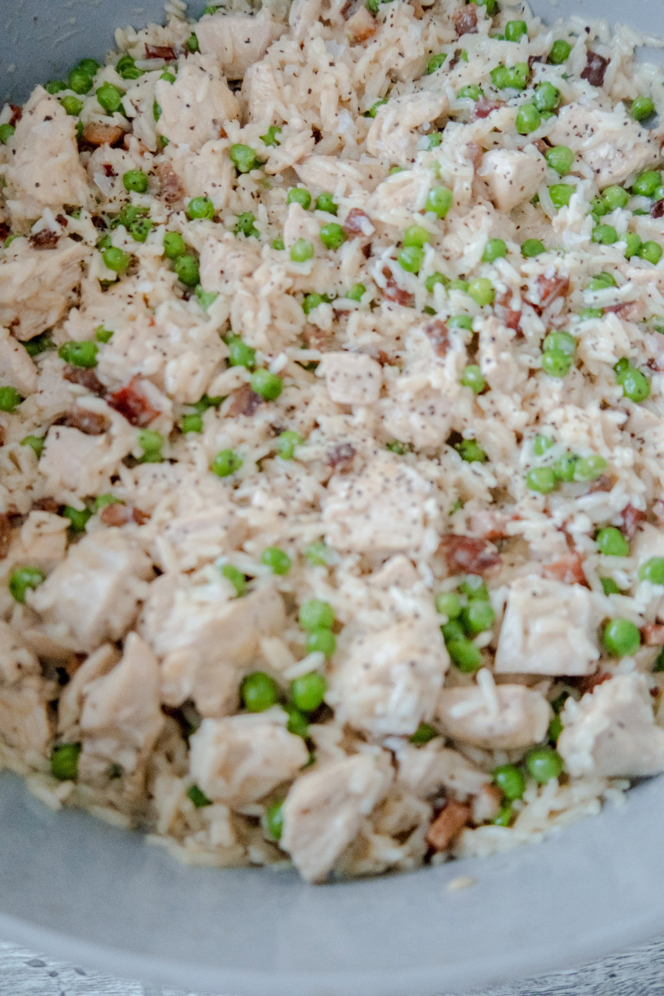 A close-up of a dish containing cooked rice, diced chicken, green peas, bits of pancetta, and a sprinkle of black pepper.