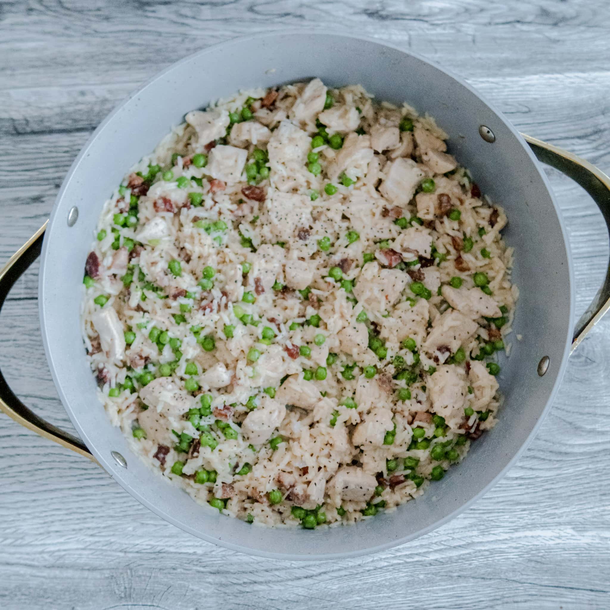An overhead shot of a large pan filled with creamy rice mixed with chunks of chicken, green peas, and pieces of bacon, placed on a light wooden surface.